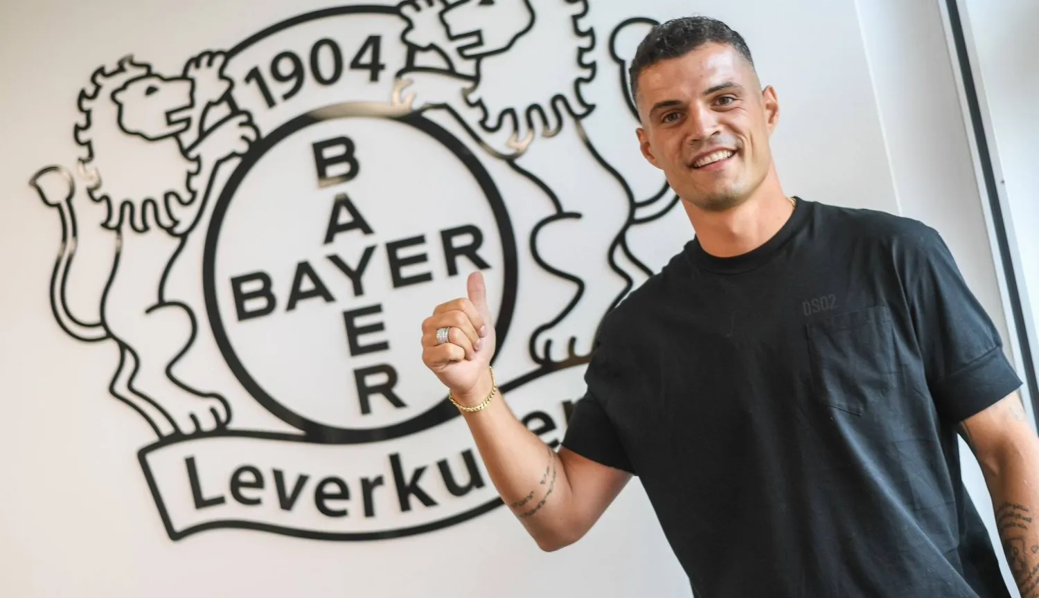 شاكا ومن خلفه شعار «باير ليفركوزن» بعد مراسم توقيع العقد (نادي باير ليفركوزن)