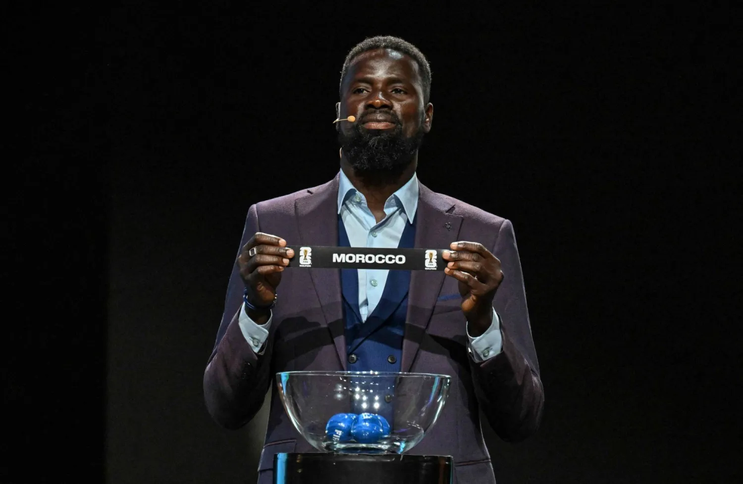 النجم المعتزل إيمانويل أبوي يرفع ورقة المغرب خلال مراسم القرعة (أ.ف.ب)