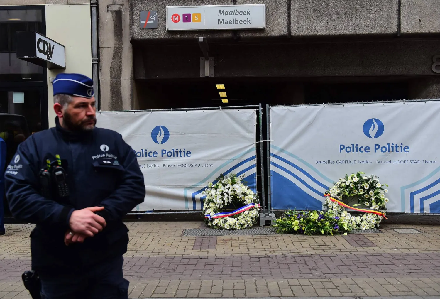 شرطي يقف بجانب أكاليل الزهور التي وضعها رئيس الوزراء الفرنسي مانويل فالس ورئيس الوزراء البلجيكي شارل ميشال ورئيس المفوضية الأوروبية جان كلود يونكر عند مدخل محطة مترو مالبيك مالبيك في بروكسل في 23 مارس 2016 بعد يوم من التفجيرات التي ضربت العاصمة البلجيكية (أ.ف.ب)