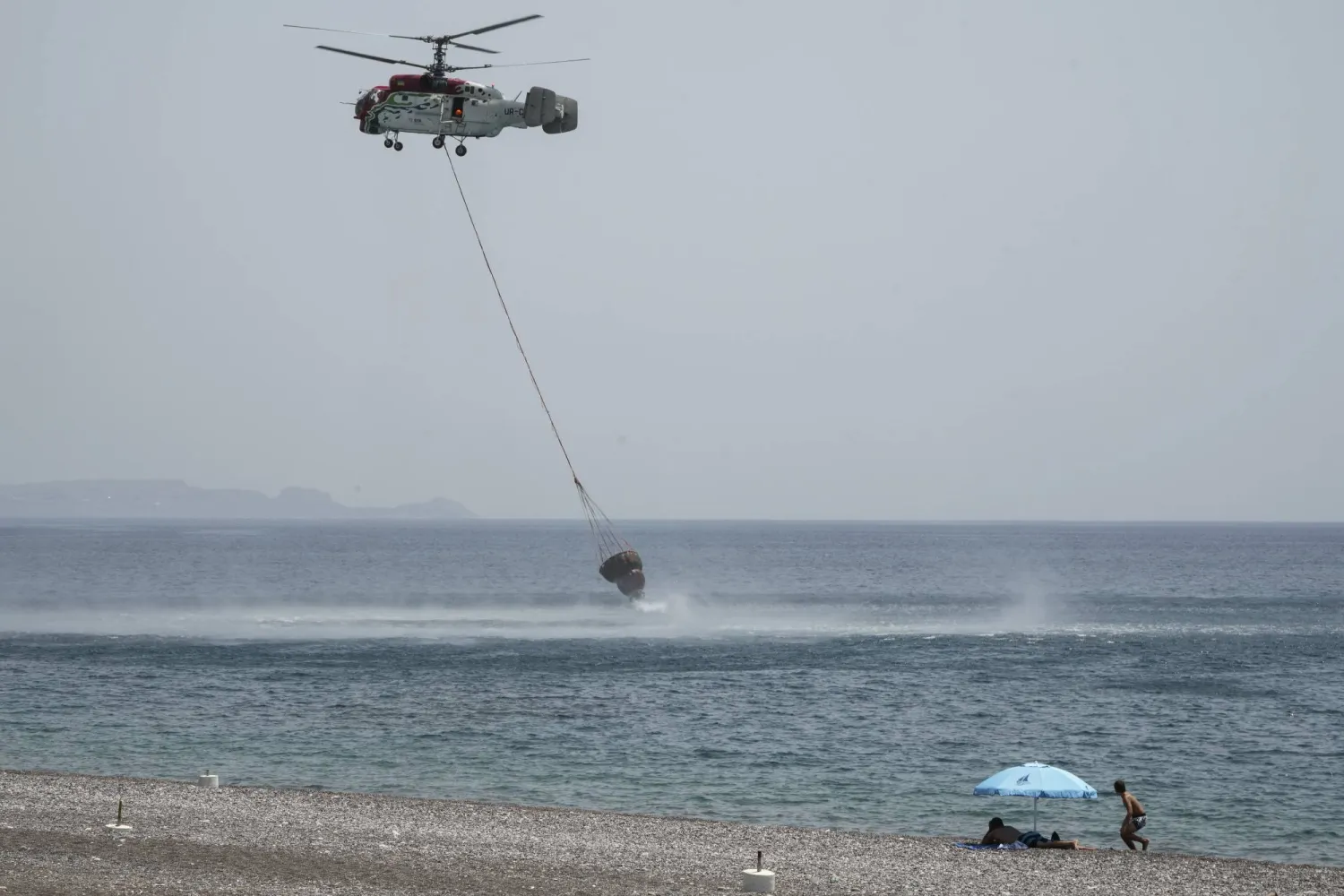 FILE - Beachgoers watch a helicopter filling water from the sea during a wildfire, near Gennadi village, on the Aegean Sea island of Rhodes, southeastern Greece, on July 27, 2023. (AP Photo/Petros Giannakouris, File)