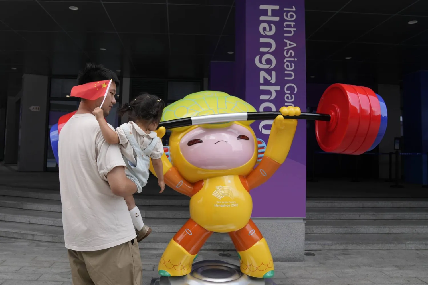 A man carries a child holding Chinese flag next to the Asian Games mascot at the weightlifting competition venue of 19th Asian Games in Hangzhou, China, Sunday, Oct. 1, 2023. (AP Photo/Aijaz Rahi)