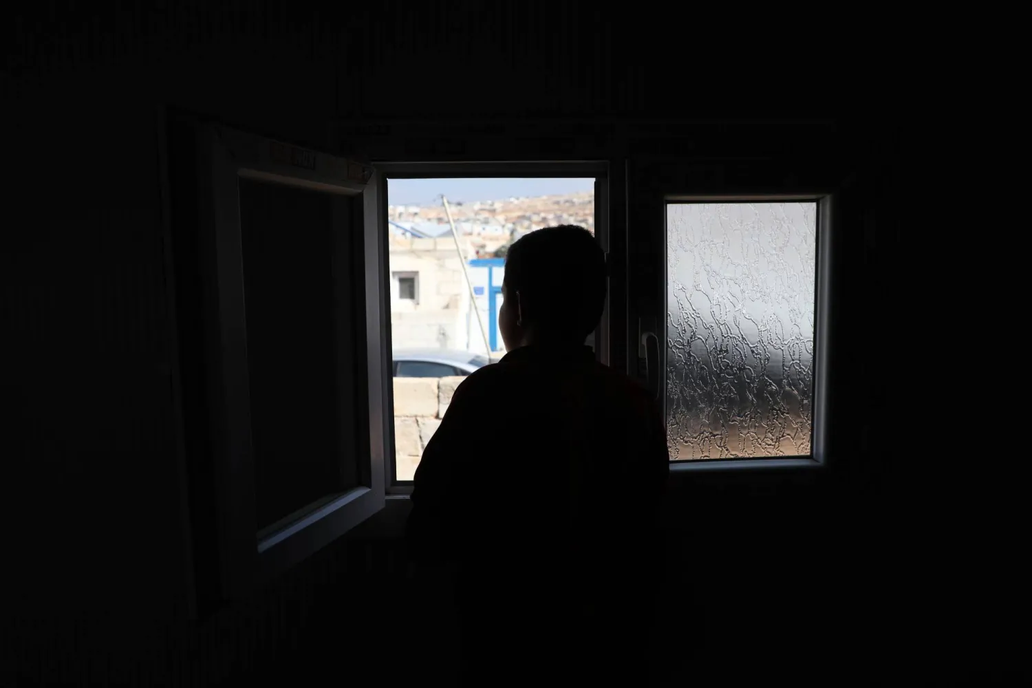 الطفل عبد السلام قرب نافذة تطل على مخيم يقيم فيه بعد نزوحه من قريته التي عاش فيها صدمة مقتل أقاربه بغارات (الشرق الأوسط)