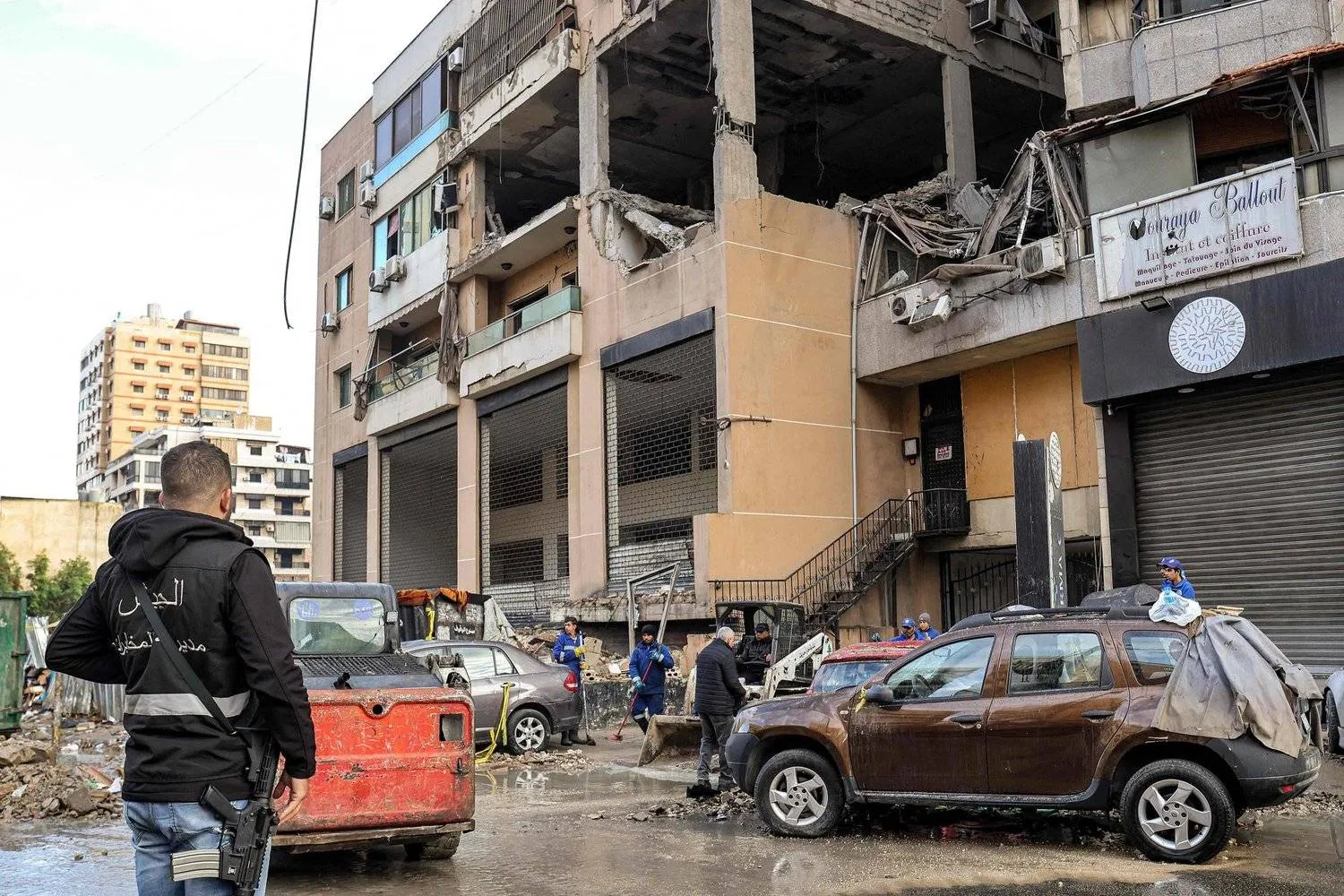 عنصر من استخبارات الجيش اللبناني بجانب عمال ينظفون موقع استهداف العاروري في الضاحية الجنوبية (أ.ف.ب)