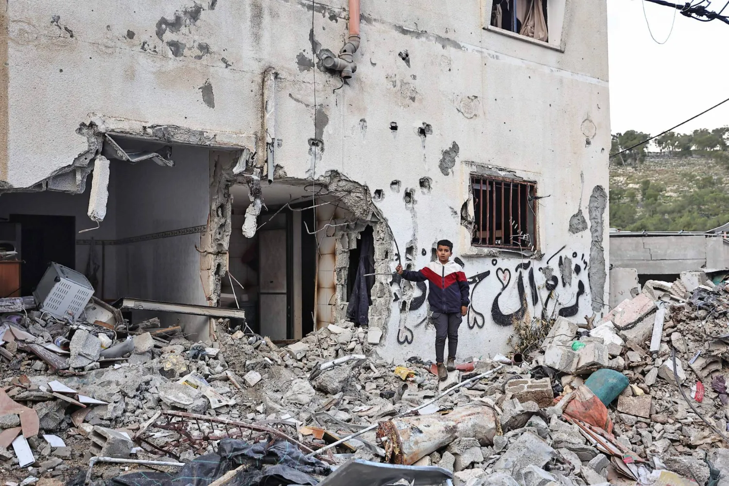 طفل يقف بجوار مبنى تضرَّر خلال غارة إسرائيلية على مخيم نور شمس للاجئين الفلسطينيين بالقرب من مدينة طولكرم بالضفة الغربية (أ.ف.ب)