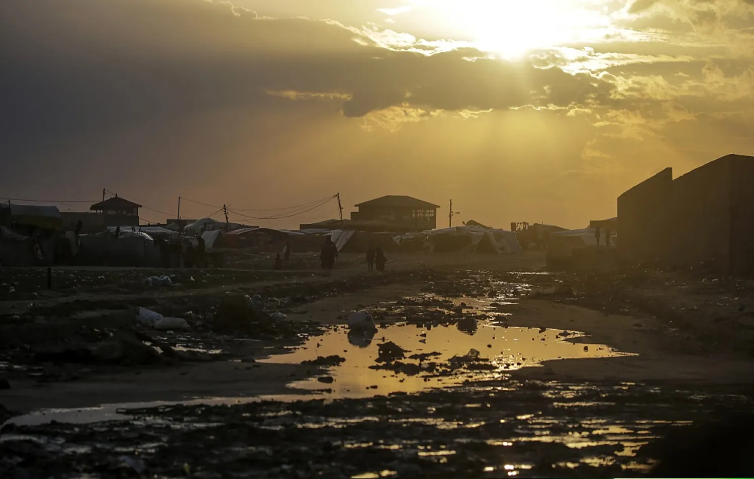 Displaced Palestinians who fled their houses from the northern Gaza Strip walk past a rain water puddle next to their shelters during a cold evening in Deir Al Balah town, southern Gaza Strip, 30 January 2024. (EPA) 