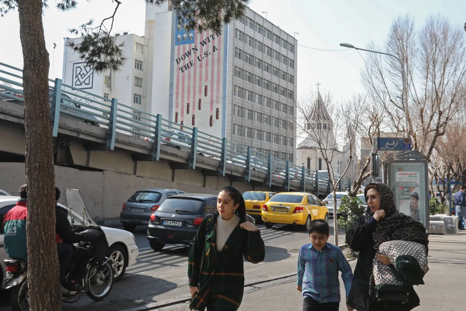 A family walks past an anti-US mural covering the wall of a building in the background in central Tehran on January 30, 2024. (Photo by ATTA KENARE / AFP)