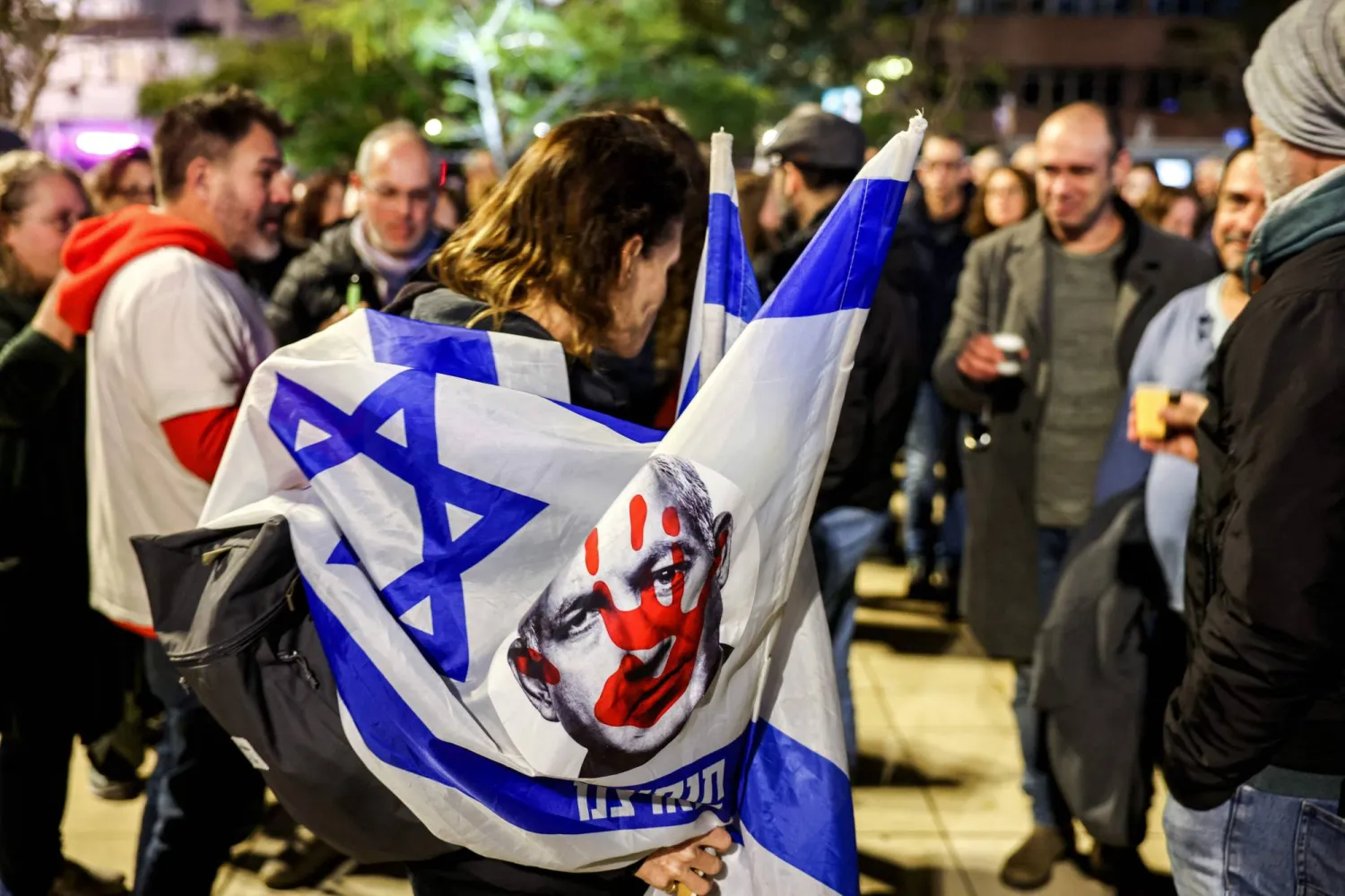 متظاهر يحمل العلم الإسرائيلي وعليه رسم لوجه رئيس الوزراء الإسرائيلي خلال مظاهرة مناهضة للحكومة في ميدان هابيما في تل أبيب  (أ.ف.ب)