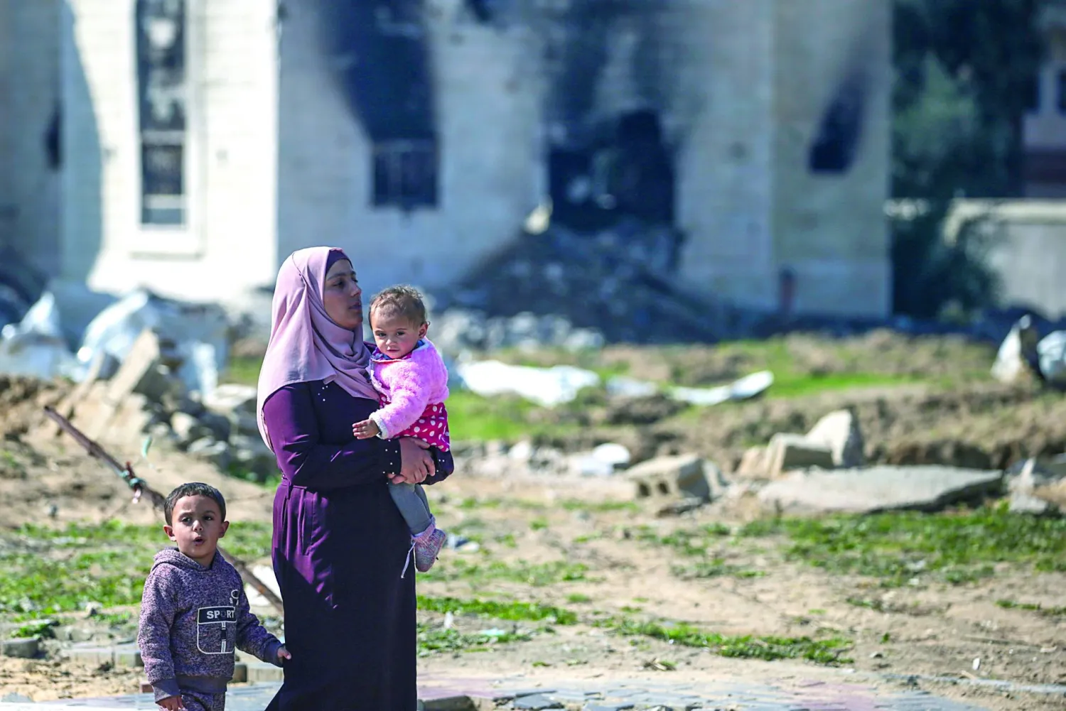 
فلسطينية مع طفليها بجوار منازل دمرتها غارات إسرائيلية على مخيم المغازي جنوب  غزة (إ.ب.أ)