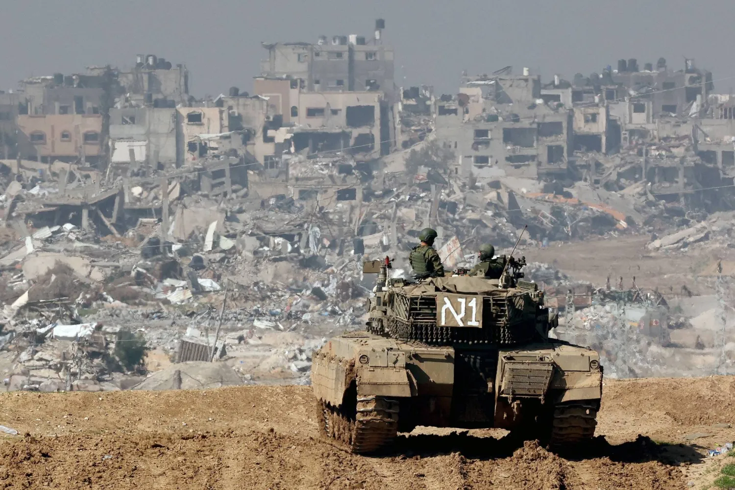 A picture taken from a position in southern Israel along the border with the Gaza Strip on January 19, 2024, shows an Israeli tank rolling along the fence as damaged buildings are seen in the Gaza strip amid continuing battles between Israel and Hamas group. (Photo by JACK GUEZ / AFP)