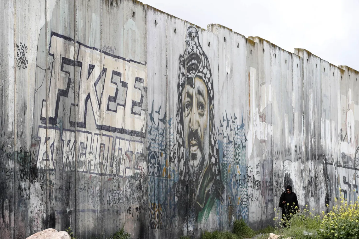 A Palestinian woman walks near a mural depicting Palestinian leader and Fatah founder Yasser Arafat painted on a section of the separation barrier between the West Bank and Israel, near the Israeli checkpoint of Qalandia, between the West Bank and Jerusalem, 15 March 2024.  EPA/ALAA BADARNEH