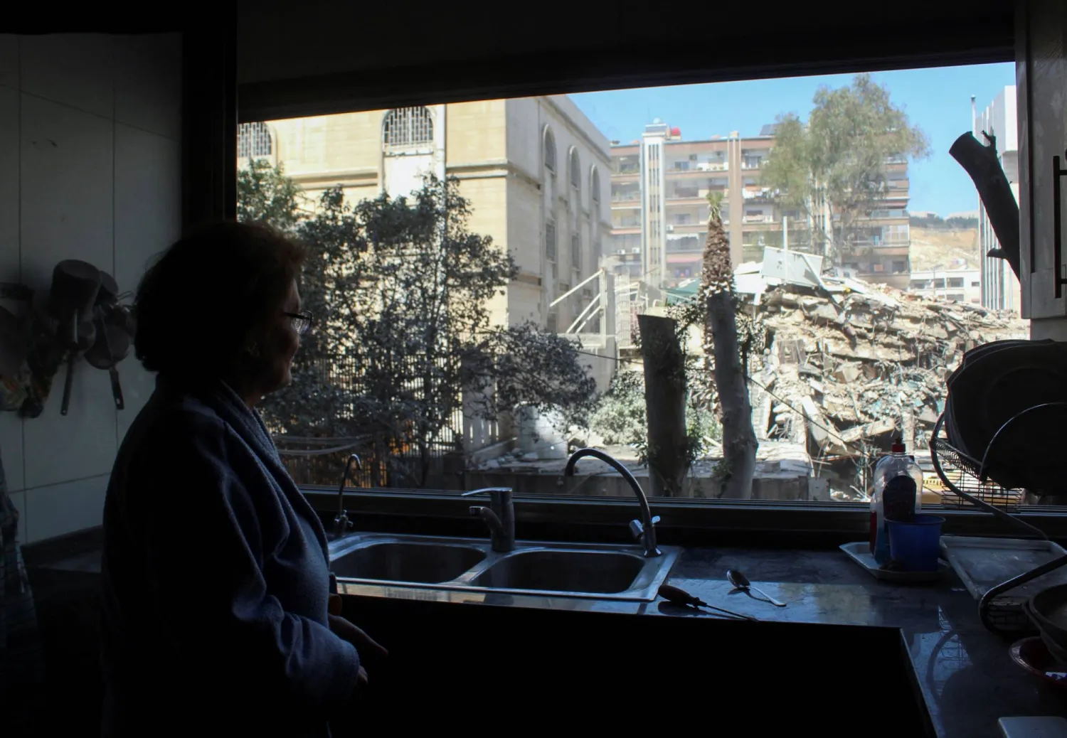سورية تراقب من مطبخ منزلها الذي يطل على القنصلية الإيرانية في دمشق ركام المبنى الذي دمرته الغارة الإسرائيلية الاثنين (رويترز)