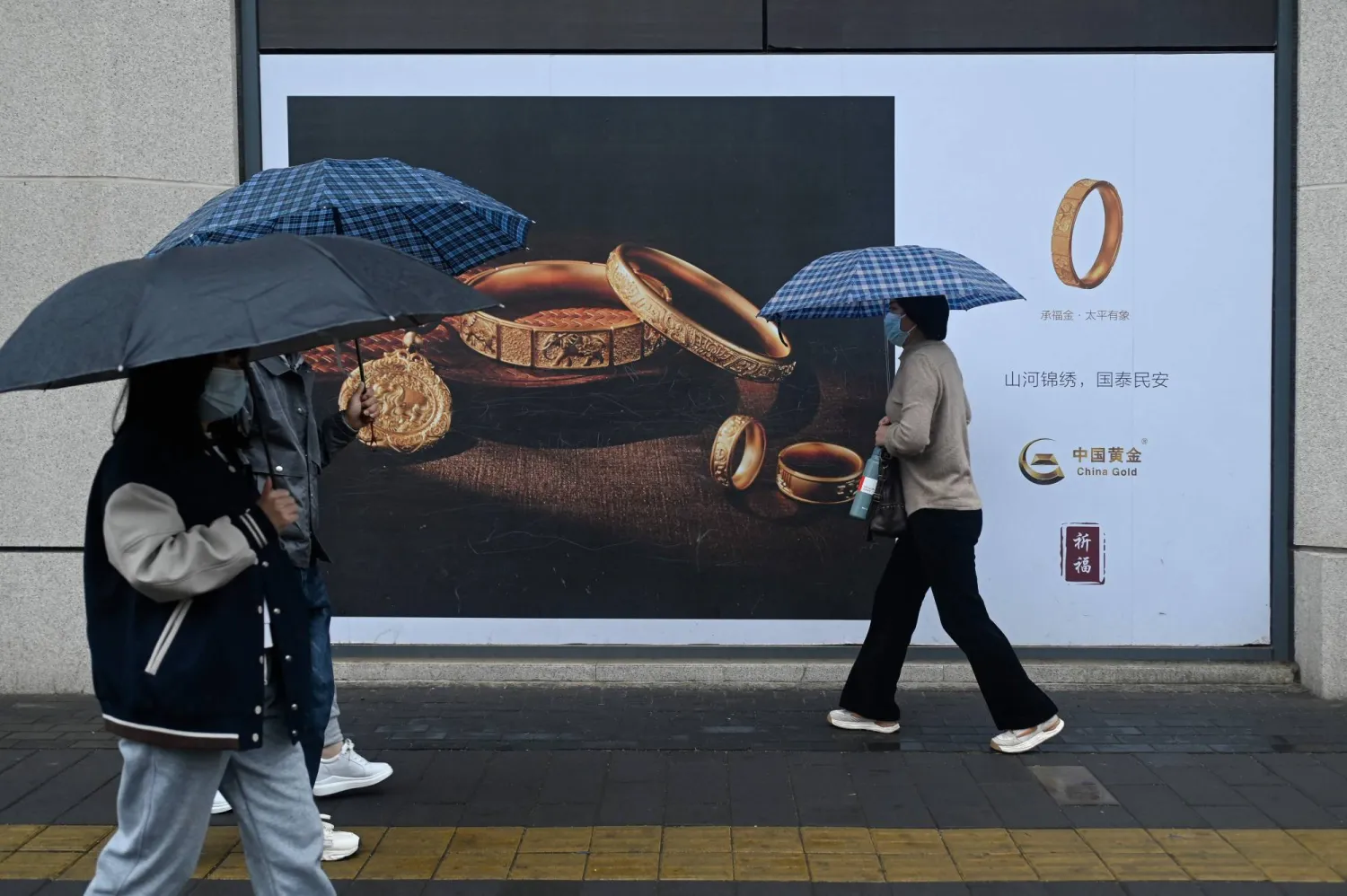 People holding umbrellas walk past a gold advertisement on a rainy day in Beijing on April 10, 2024. (Photo by WANG Zhao / AFP)