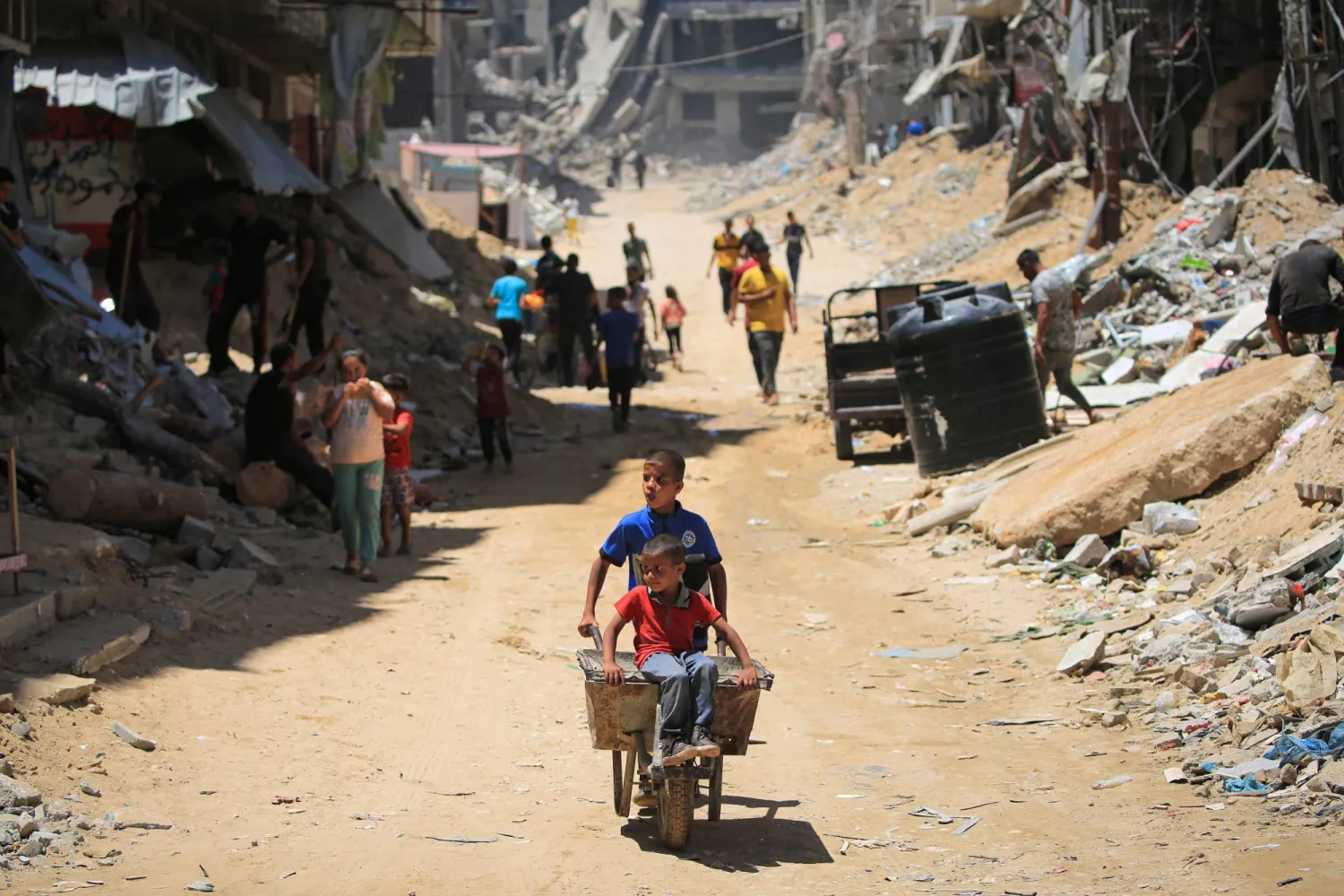A Palestinian child pushes another child sitting in a wheelbarrow along a narrow street past destroyed buildings in Khan Yunis, in the southern Gaza Strip on June 11, 2024. (Photo by Eyad BABA / AFP)