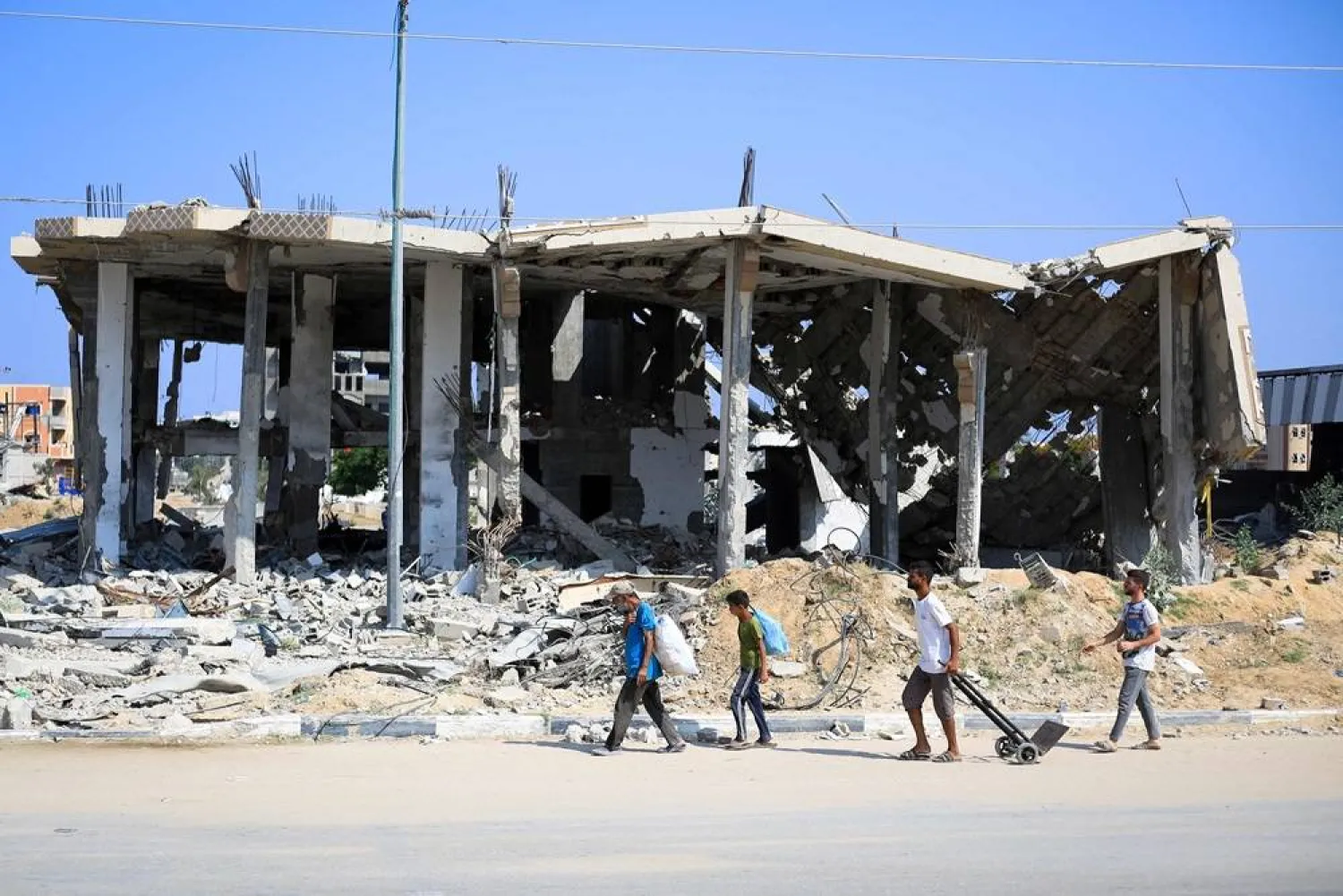 Displaced Palestinians carry belongings as they walk in front of a destroyed building in Deir el-Balah in the central Gaza Strip on July 7, 2024. (AFP)