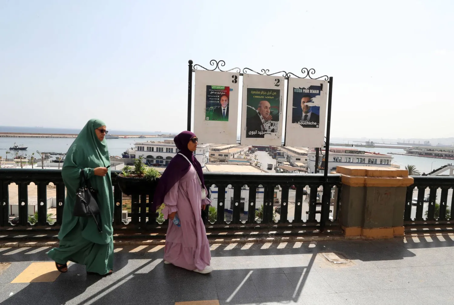 جزائريتان تمران بجانب لوحة دعائية تحمل صور المرشحين الثلاثة (إ.ب.أ)