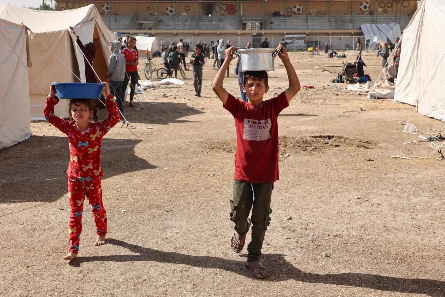 srail ordusunun kuzey Gazze Şeridi'ndeki operasyonlarından ailesiyle birlikte kaçan yerinden edilmiş Filistinli bir çocuk, "Yermuk Stadyumu"nda yeni kurulan çadırların önünde yemek takımlarını taşıyor (AFP)