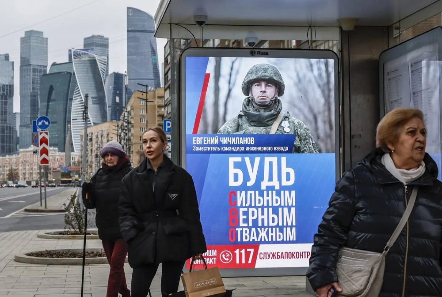 People wait at a bus stop with electronic screen showing an advertisement image depicting Russian soldier and the slogan "Be strong, faithful, courageous" in front of towers of business center Moscow-City in Moscow, Russia, 14 November 2024. (EPA) 