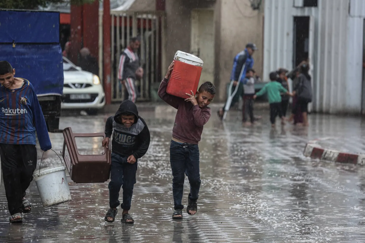 أطفال فلسطينيون يحملون بعض الأغراض ويركضون في المكان بينما يبدأ المطر بالهطول في دير البلح (د.ب.أ)