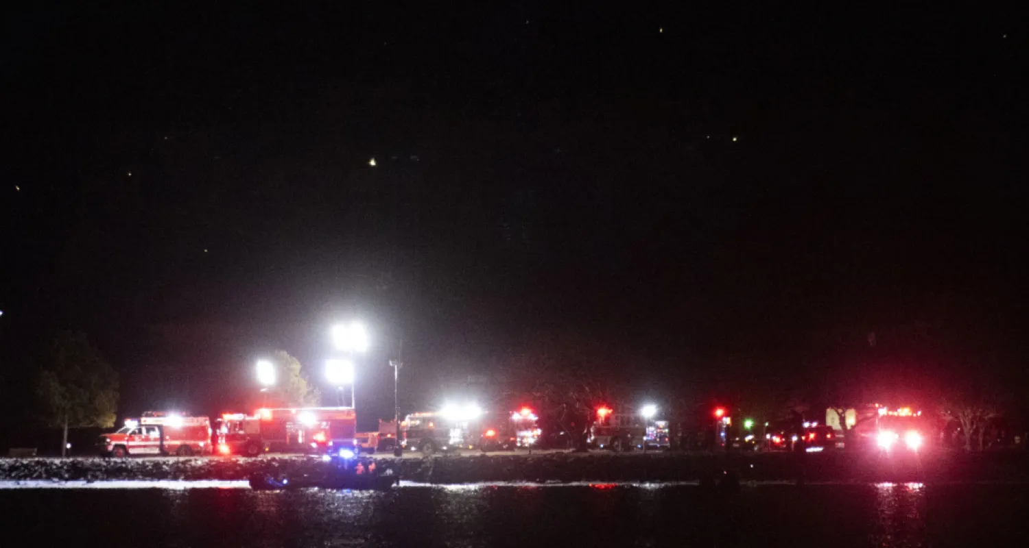 A boat on the Potomac River, cruises past emergency response vehicles seen staging at Joint Base Anacostia Bolling, in the early morning hour, Thursday, Jan. 30, 2025 in Washington, as seen from across the river near Ronald Reagan Washington National Airport in Arlington, Va. (AP Photo/Kevin Wolf)