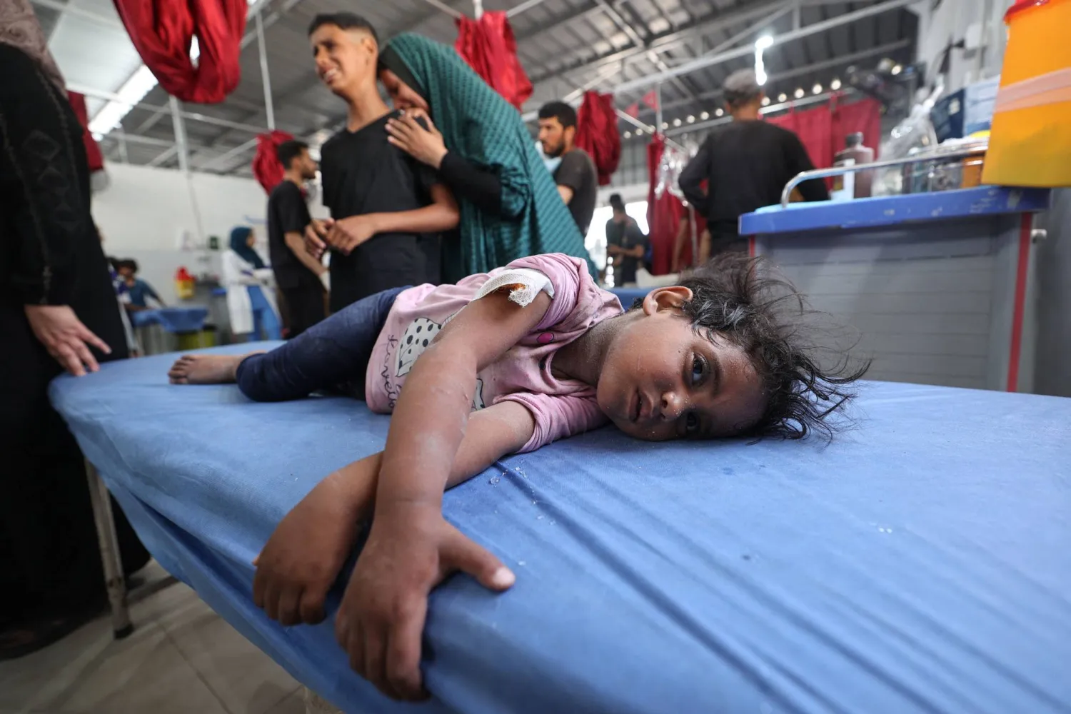 امرأة وطفل يبكيان بينما ترقد طفلة صغيرة على سرير مستشفى تنتظر الإسعافات الأولية في مستشفى العودة بمخيم النصيرات للاجئين وسط قطاع غزة (أ.ف.ب)