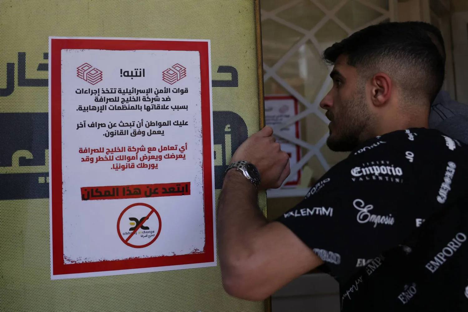 رجل يقف بجانب لافتة علّقتها قوات الاحتلال الإسرائيلية على جدران محل صرافة في الخليل تحذر من التعامل معه بمداهمات جرت يوم الثلاثاء (أ.ف.ب)