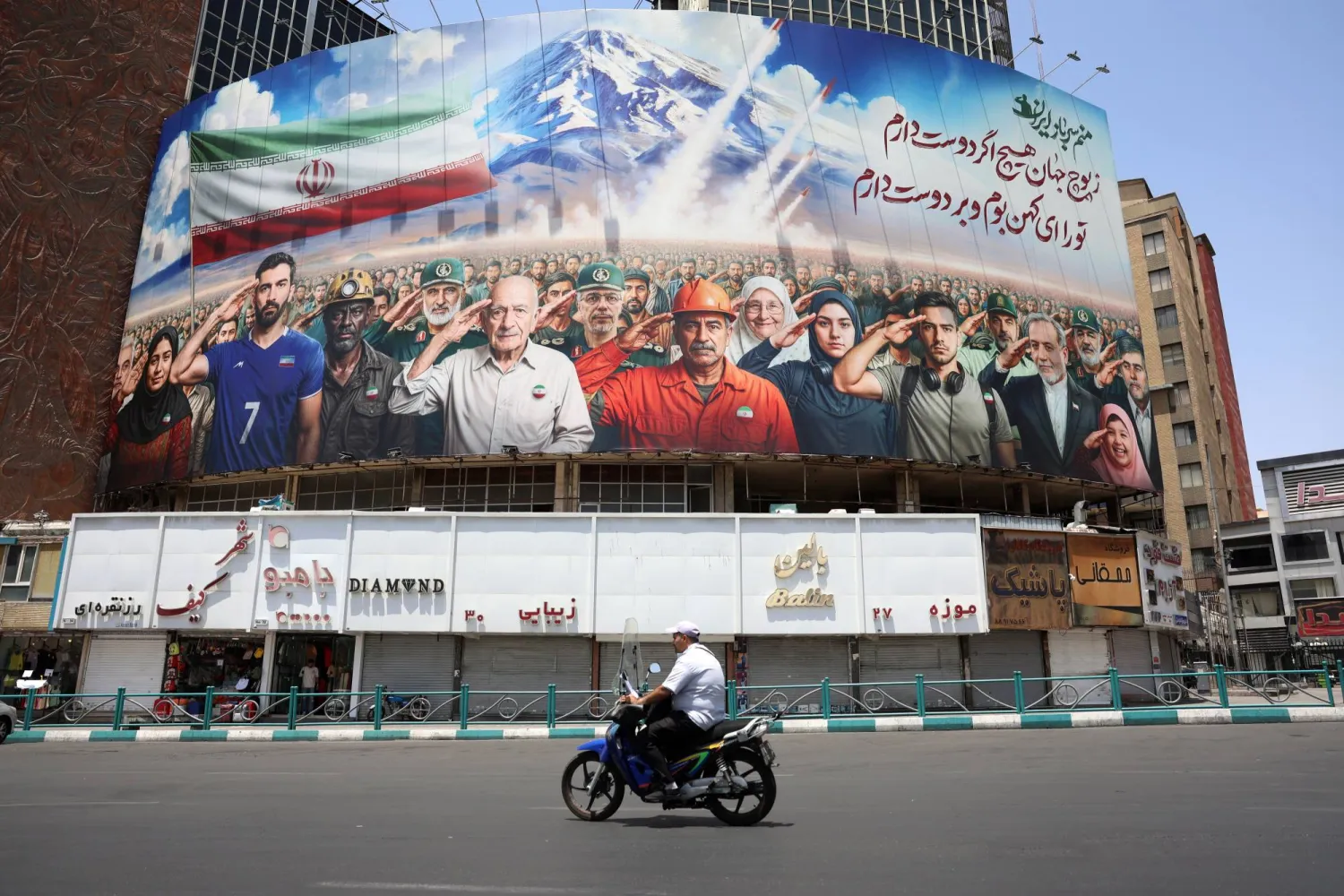 رجل إيراني يقود دراجة نارية أمام لوحة إعلانية تُظهر عدداً من الإيرانيين من بينهم عسكريون ووزير الخارجية الإيراني عباس عراقجي (الثالث يمين) يؤدون التحية العسكرية ورسالة كُتب عليها: «كلنا جنود إيران» في ساحة «ولي عصر» بطهران (إ.ب.أ)