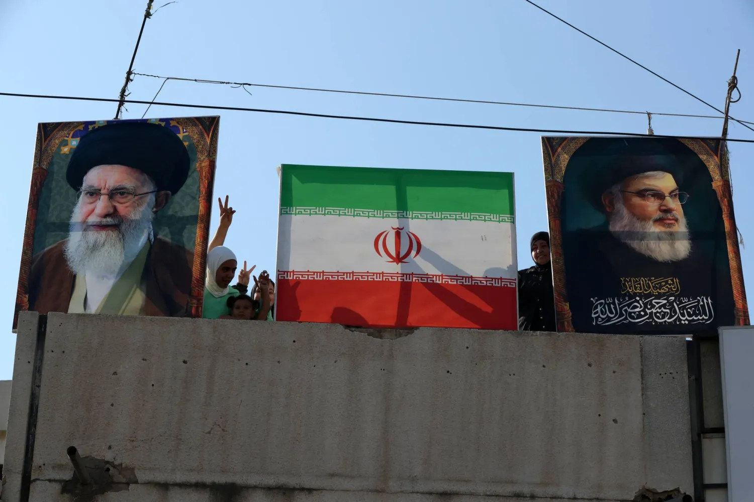 أشخاص يرفعون علامة النصر بجوار صورة خامنئي والعلم الإيراني وصورة الراحل نصر الله (إ.ب.أ)