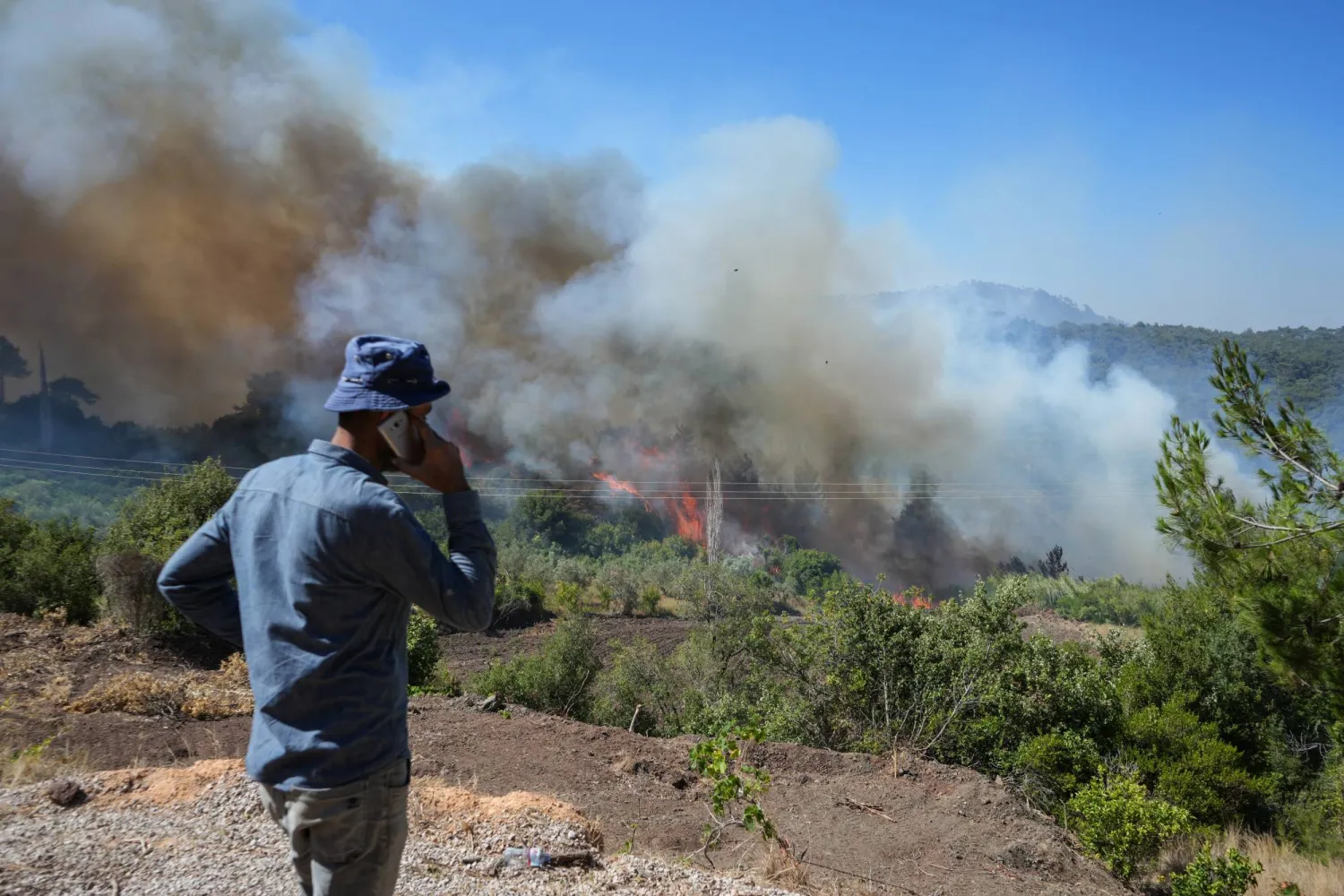 رجل يقف بالقرب من دخان يتصاعد من حريق هائل في منطقة قسطل معاف بريف اللاذقية في سوريا (إ.ب.أ)