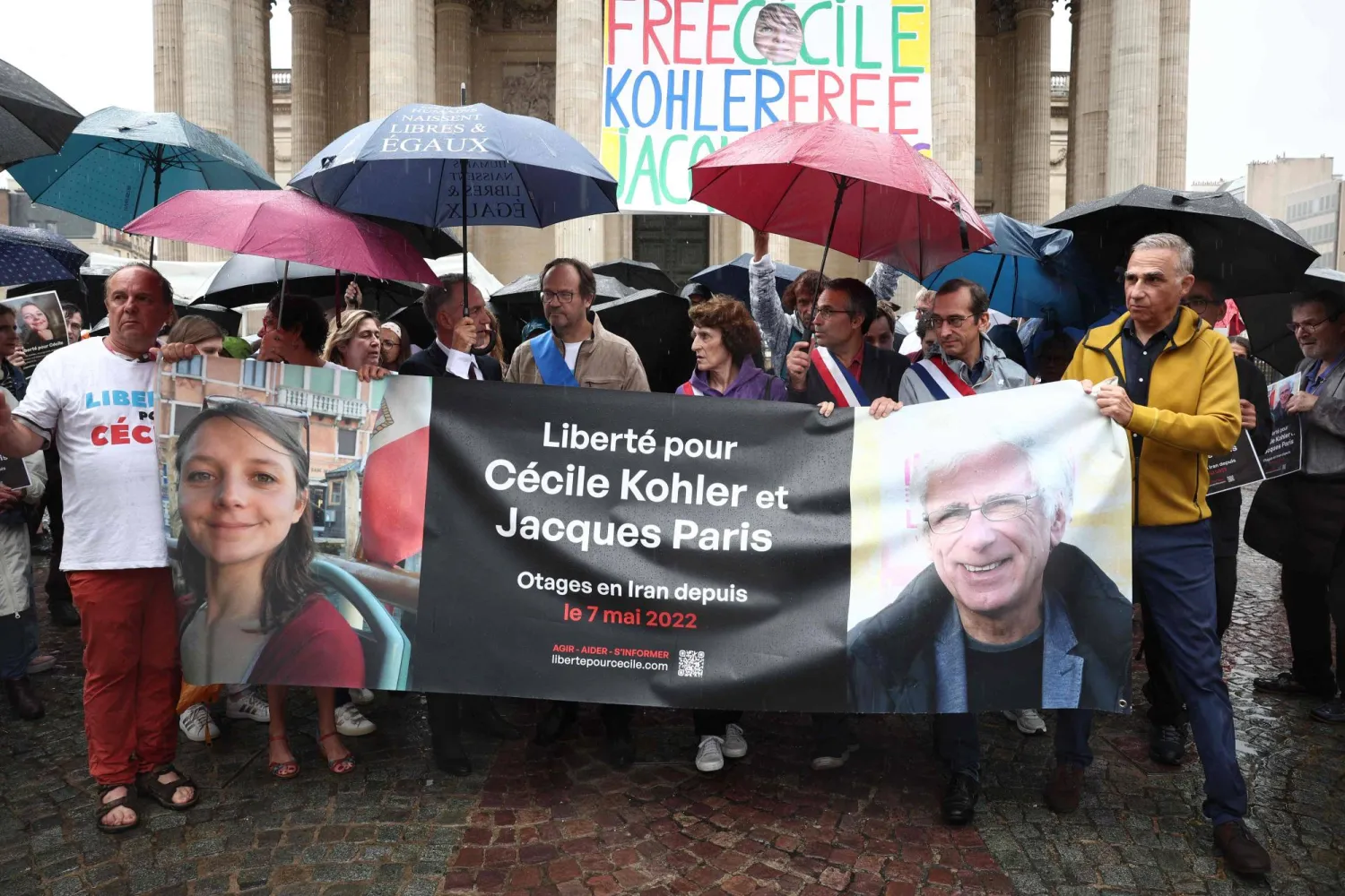Relatives and supporters pose behind a banner with the portrait of Cecile Kohler (L) and Jacaues Paris (R) who are being held in Iran since May 2022 on espionage charges, at the end of a rally in support to them at Place du Pantheon in Paris, on July 6, 2025. (Photo by Sebastien DUPUY / AFP)
