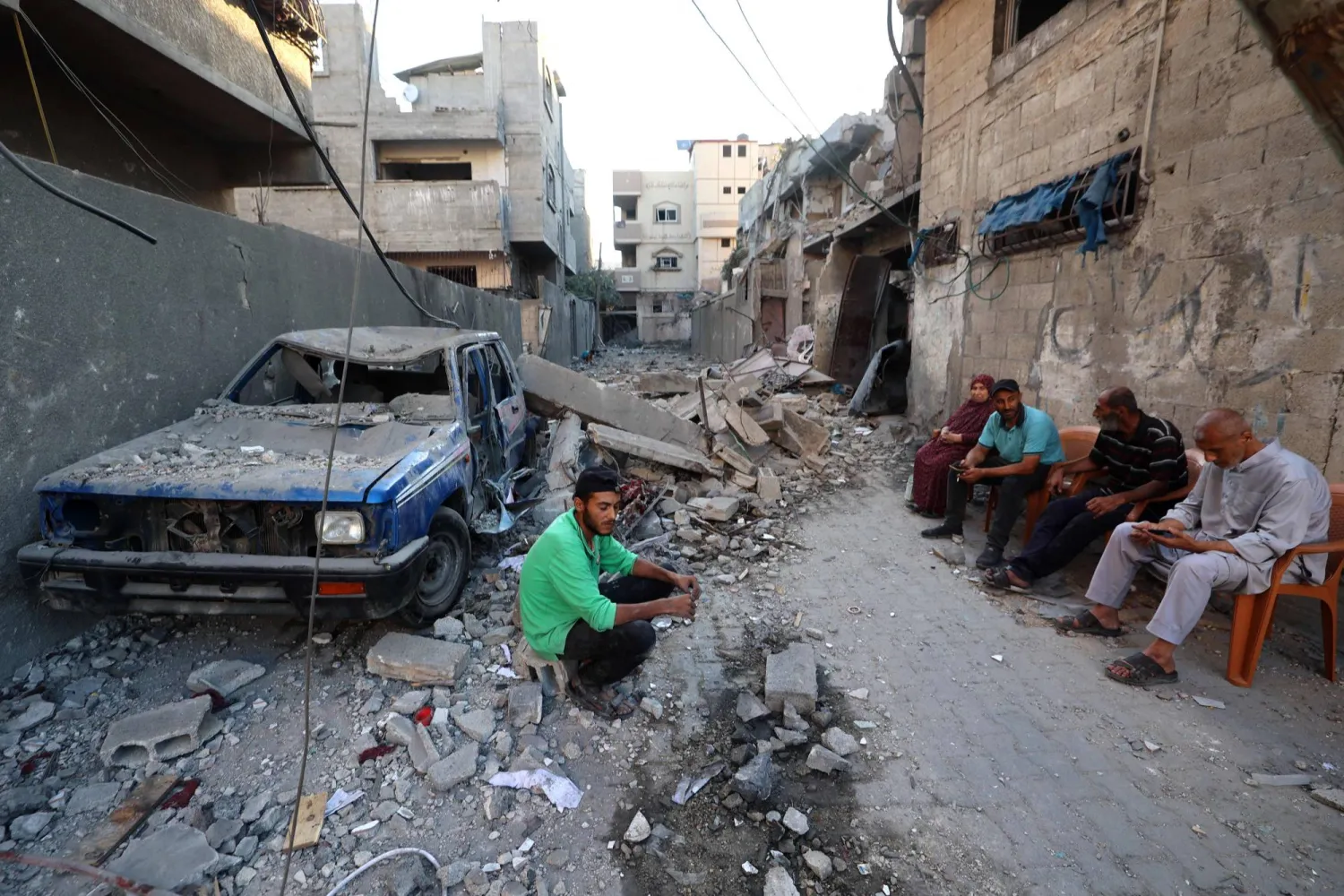 فلسطينيون يجلسون بجوار منزل مهدّم وسيارة مدمّرة جراء القصف الإسرائيلي على مخيم البريج وسط قطاع غزة (أ.ف.ب)