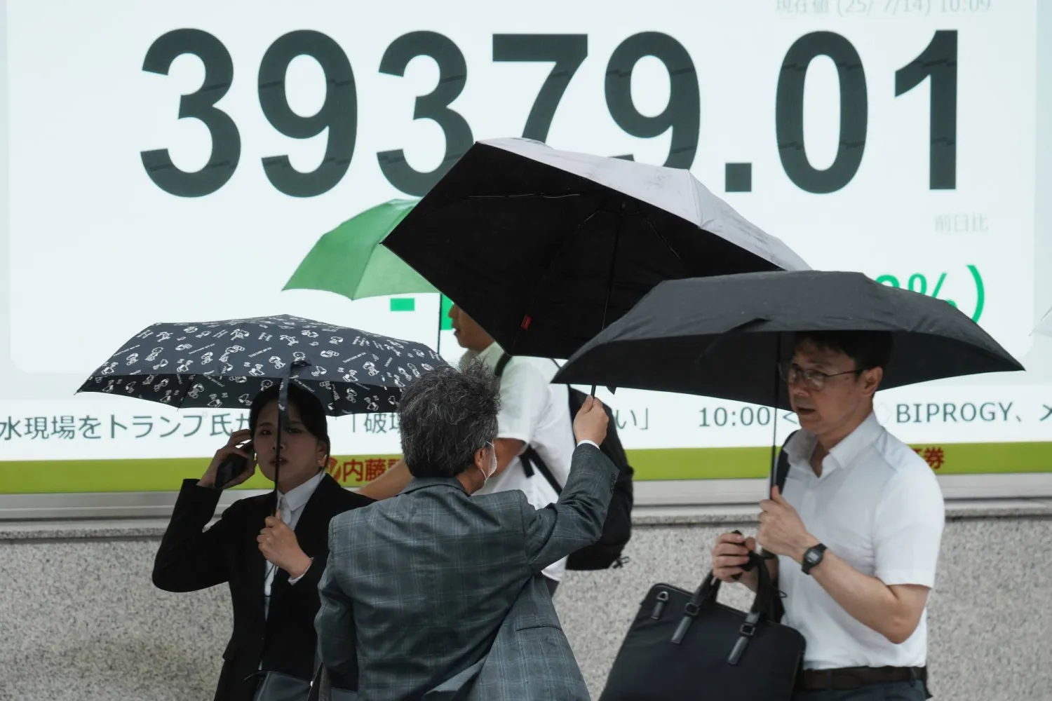 مشاة في أحد شوارع العاصمة اليابانية طوكيو يحتمون بالمظلات من الأمطار بينما تبدو خلفهم لوحة إلكترونية تظهر حركة الأسهم (أ.ب)