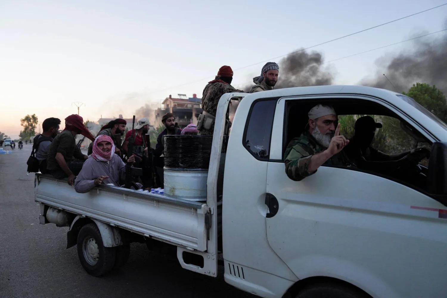 Smoke rises in the background as Bedouin fighters sit in the back of a truck, at Sweida governorate, Syria, July 18, 2025. REUTERS/Karam al-Masri