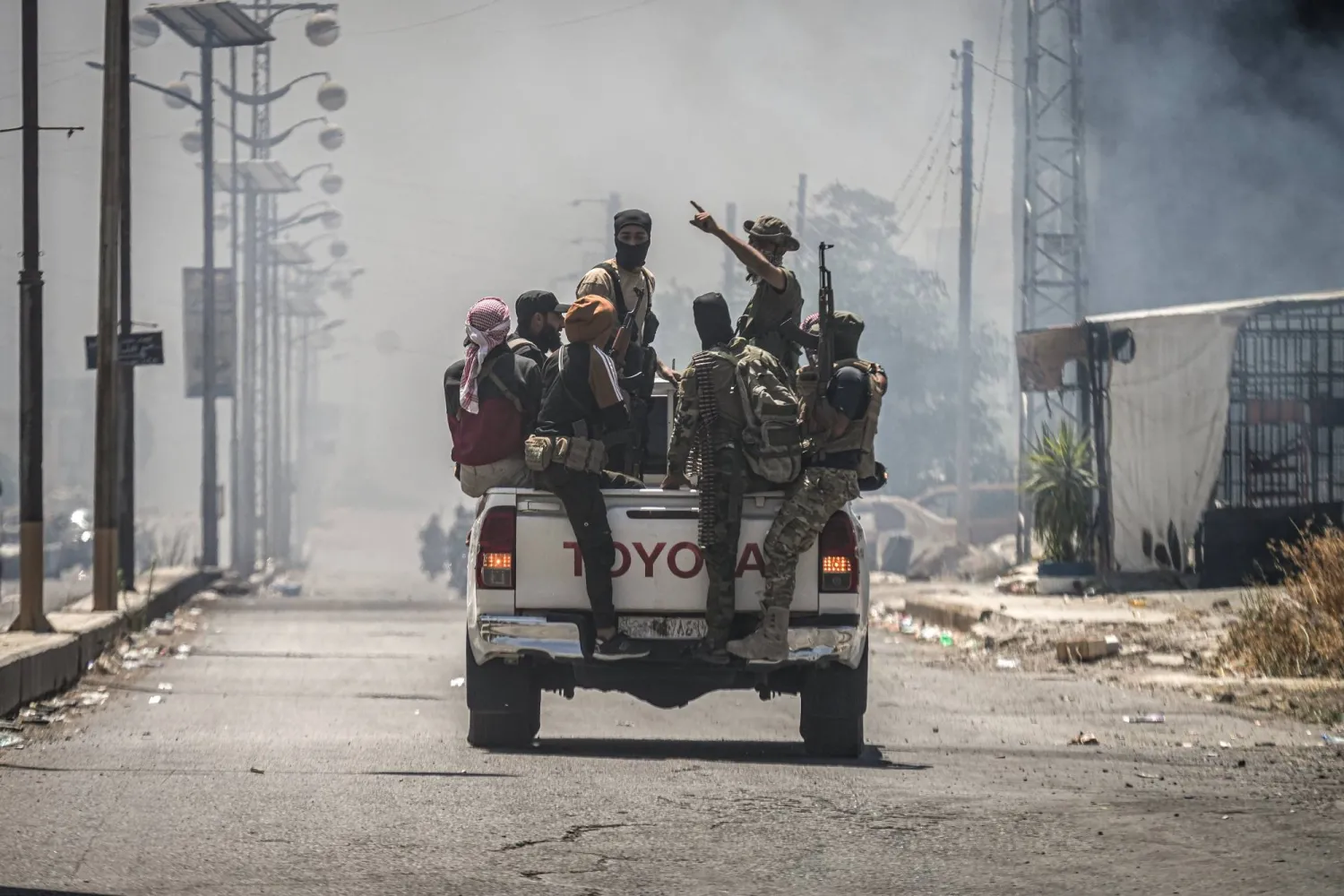 تجمّعَ مقاتلون من البدو والعشائر في سيارة بمدينة السويداء بينما يتصاعد الدخان من منازل محترقة وسط اشتباكات بين مقاتلين من العشائر وفصائل درزية محلية جنوب سوريا (د.ب.أ)