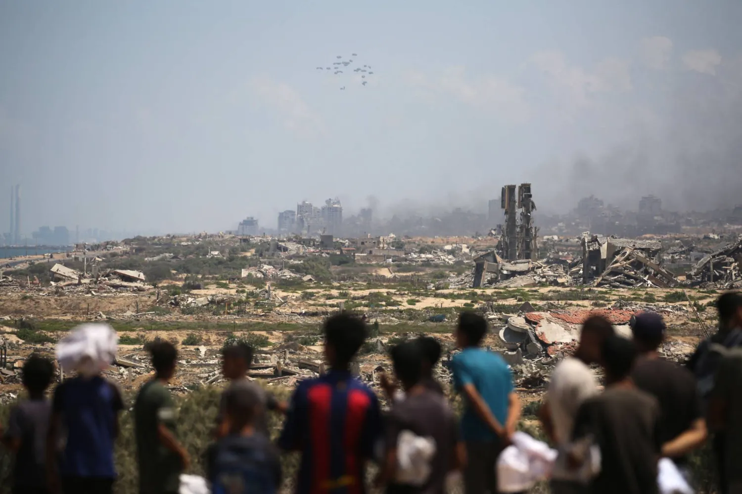 Palestinians watch aid pallets parachute down after being dropped from a military plane over Nuseirat in the central Gaza Strip during an airdrop mission above the Israel-besieged Palestinian territory on August 5, 2025. (Photo by Eyad BABA / AFP)