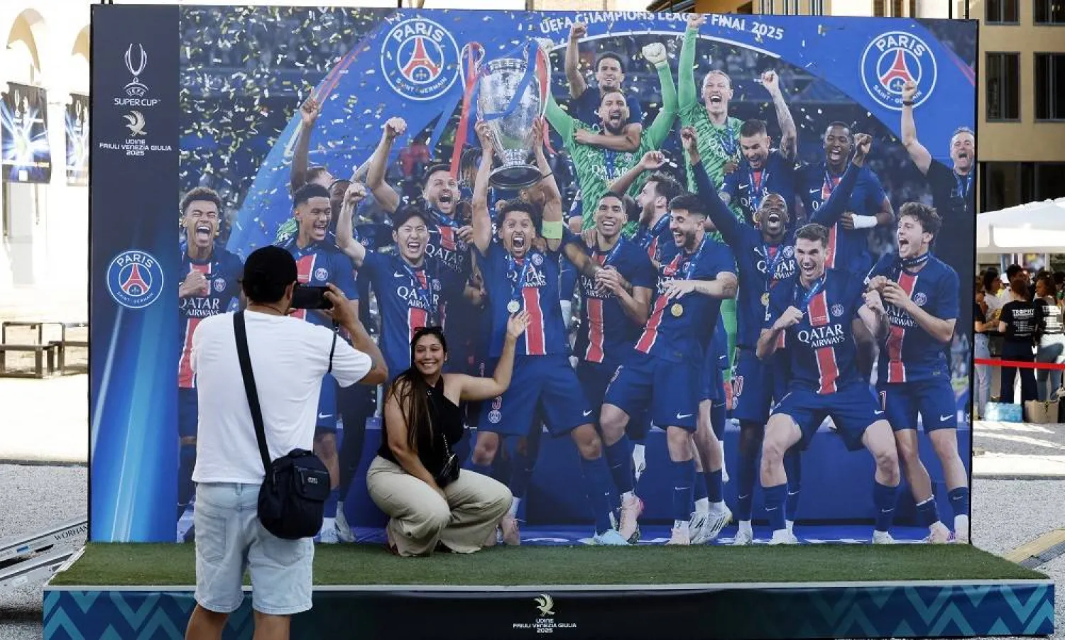 Football - UEFA Super Cup - Final - Paris St Germain v Tottenham Hotspur - Preview - Udine, Italy - August 12, 2025. People pose for photos in front of an image of Paris St Germain lifting the trophy after they won the Champions League. (Reuters) 