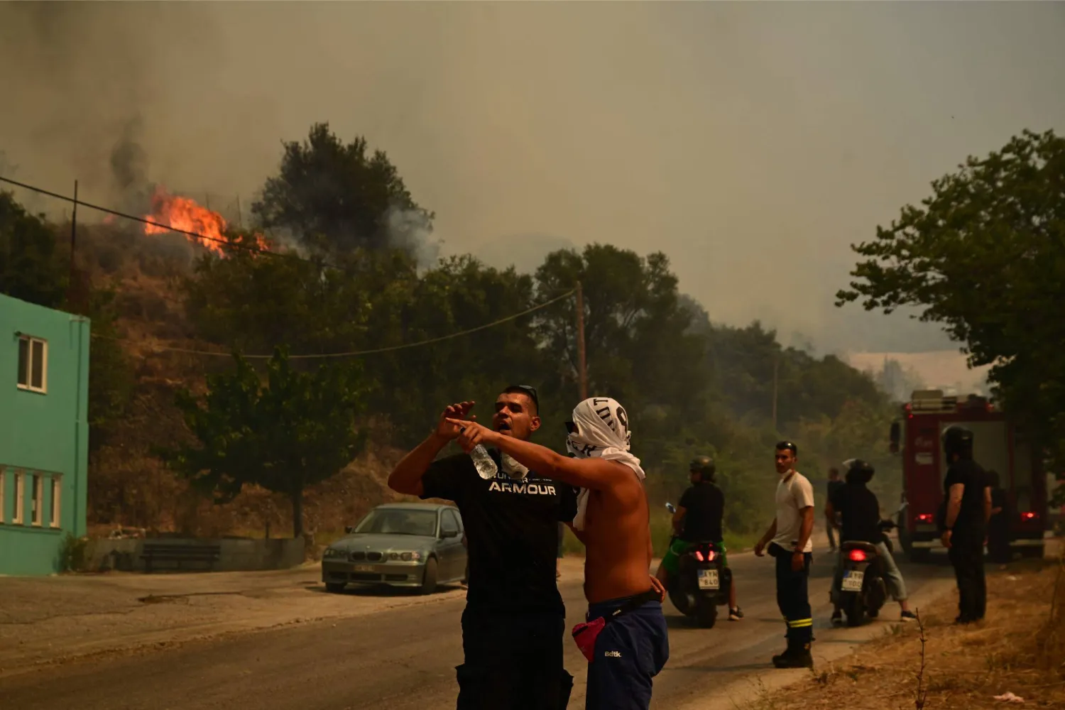 Local residents and firefighters stand along a road during efforts to extinguish a wildfire near the city of Patras, western Greece on August 13, 2025. (Photo by Aris MESSINIS / AFP)