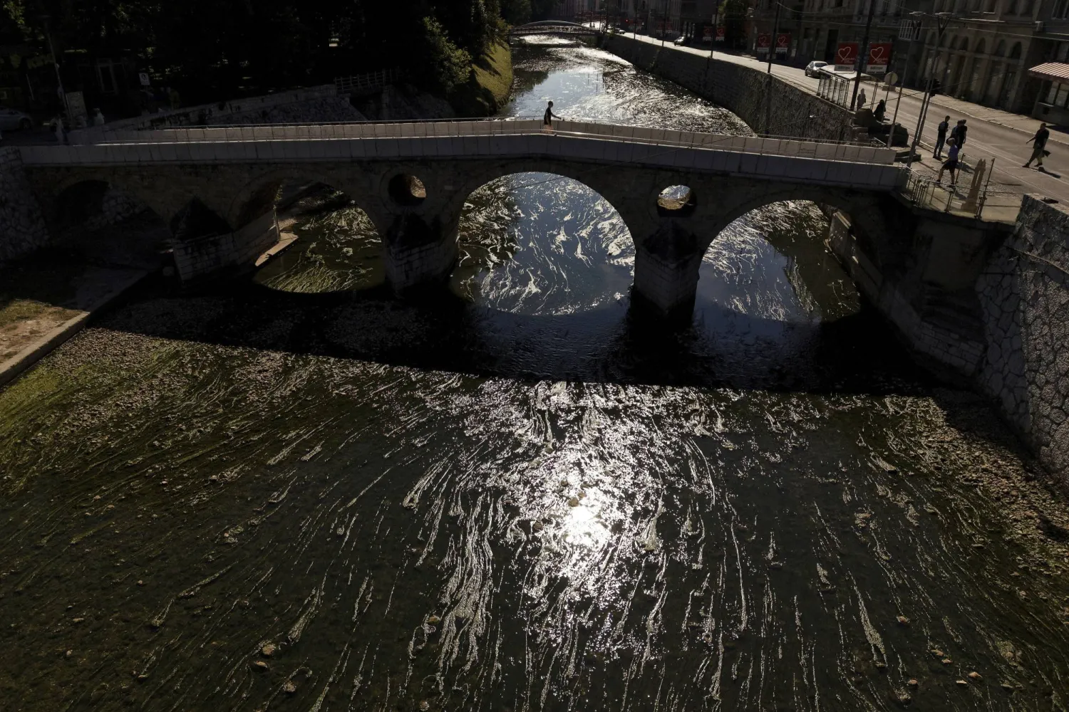 A pedestrian walks across a bridge over the Miljacka River, nearly dried up and covered with algae, amid a heatwave and drought in Sarajevo, Bosnia, Aug. 10, 2025. (AP Photo/Armin Durgut)