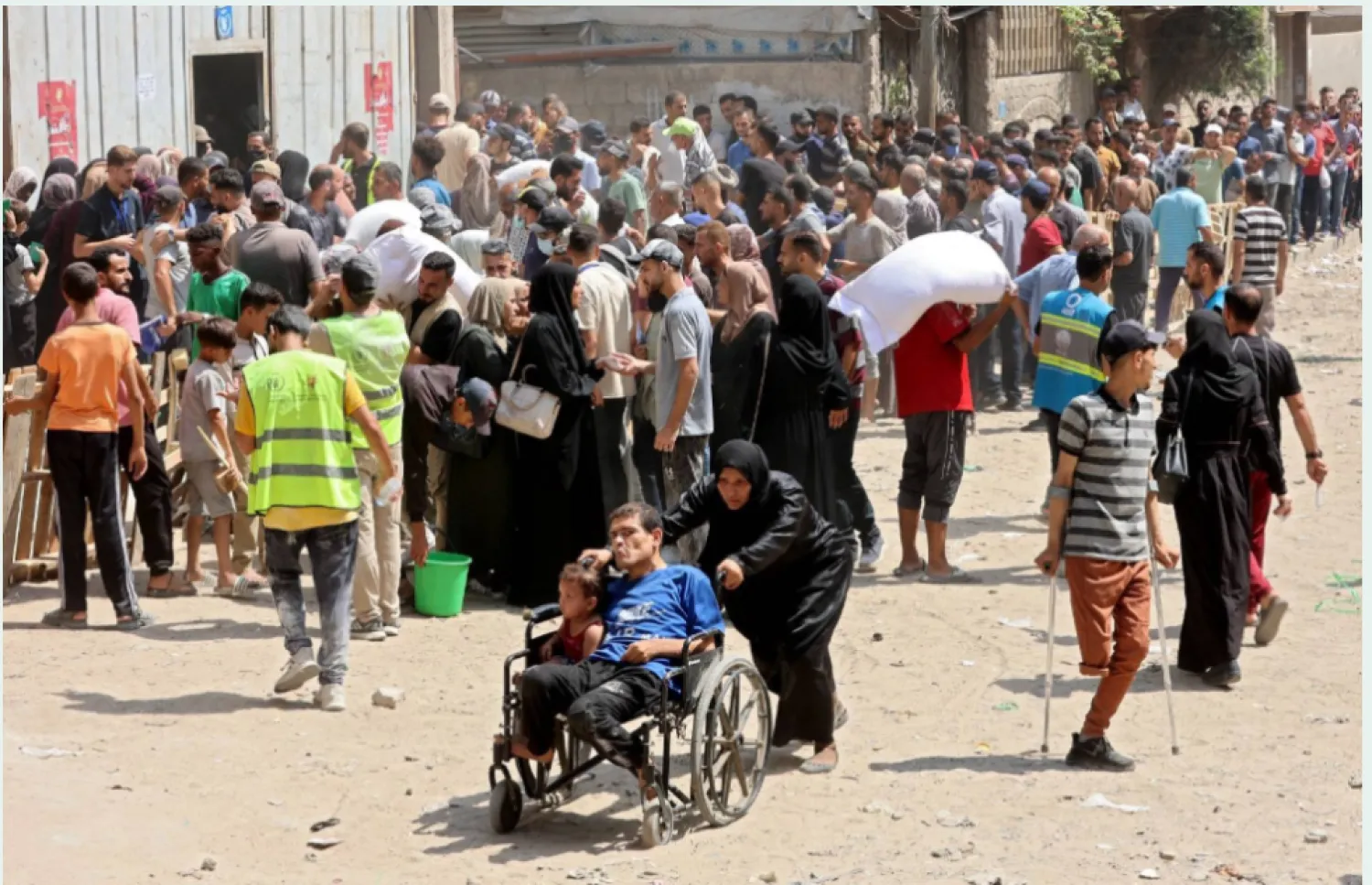 امرأة فلسطينية تدفع رجلاً وطفلاً على كرسي متحرك في حين يتجمع الناس لتلقي المساعدات (أ.ف.ب)