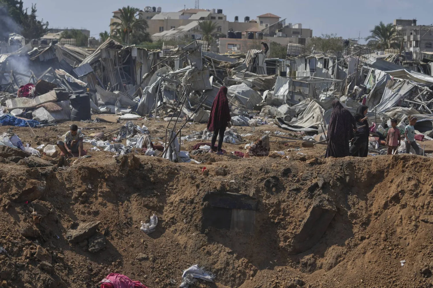 Palestinians stand on the edge of a crater after Israeli military strikes in a tent camp for displaced people near Al-Aqsa Hospital, in Deir al-Balah, Thursday, Aug. 21, 2025. (AP Photo/Jehad Alshrafi)
