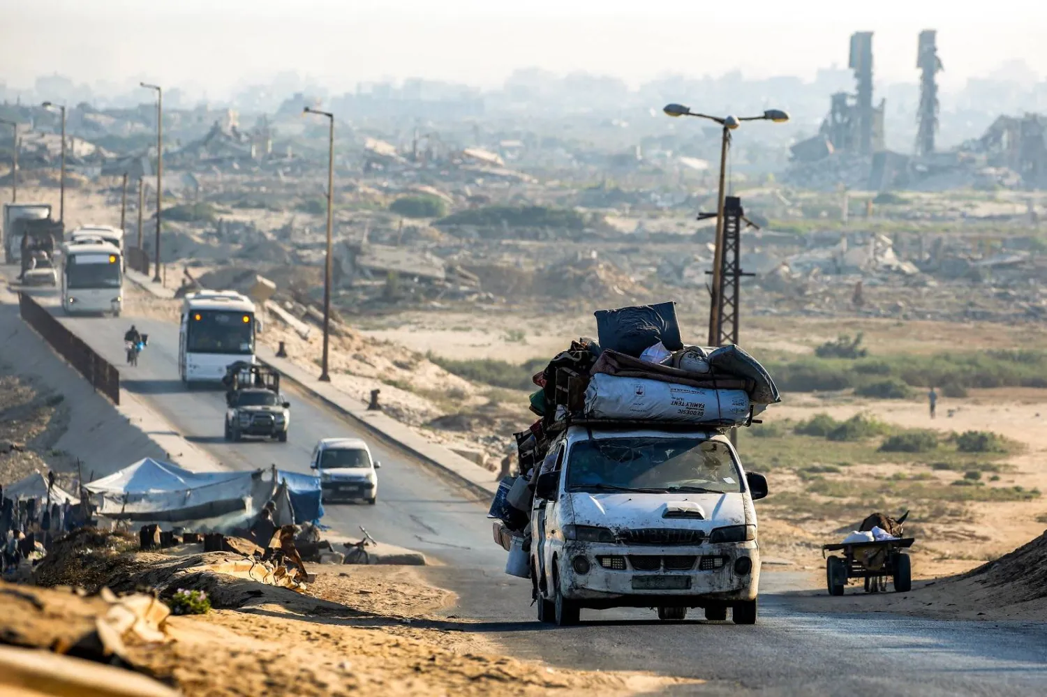  مركبات محملة بأغراض تمر بجانب مبانٍ مدمرة على طول الطريق الساحلي عبر مخيم النصيرات للاجئين الفلسطينيين (أ.ف.ب)