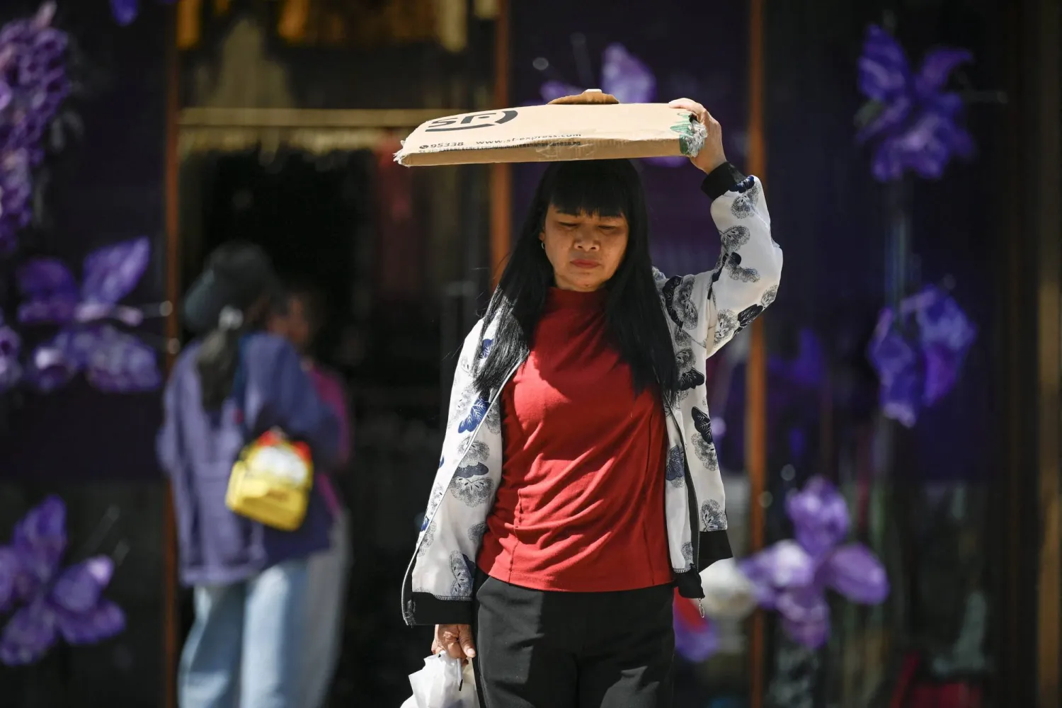 (FILES) A woman uses a piece of cardboard to shelter from the sun while walking out of a building on a hot day in Beijing on June 6, 2025. (Photo by WANG Zhao / AFP)