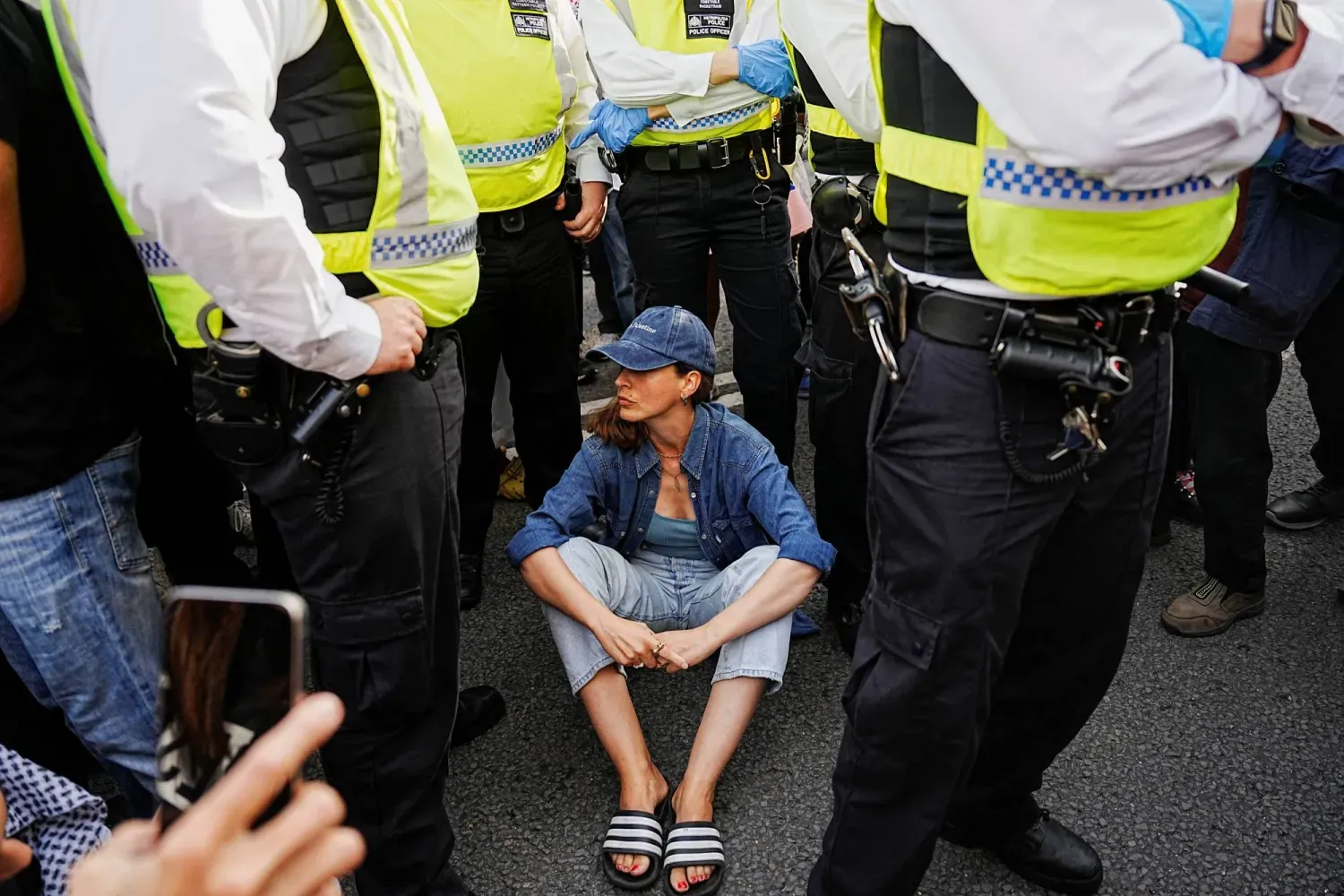 Londra'da Palestine Action grubuna eylem yasağının kaldırılması için düzenlenen bir gösteride, bir kadın İngiliz polisi tarafından çevrelenmiş halde yere otururken (Reuters)