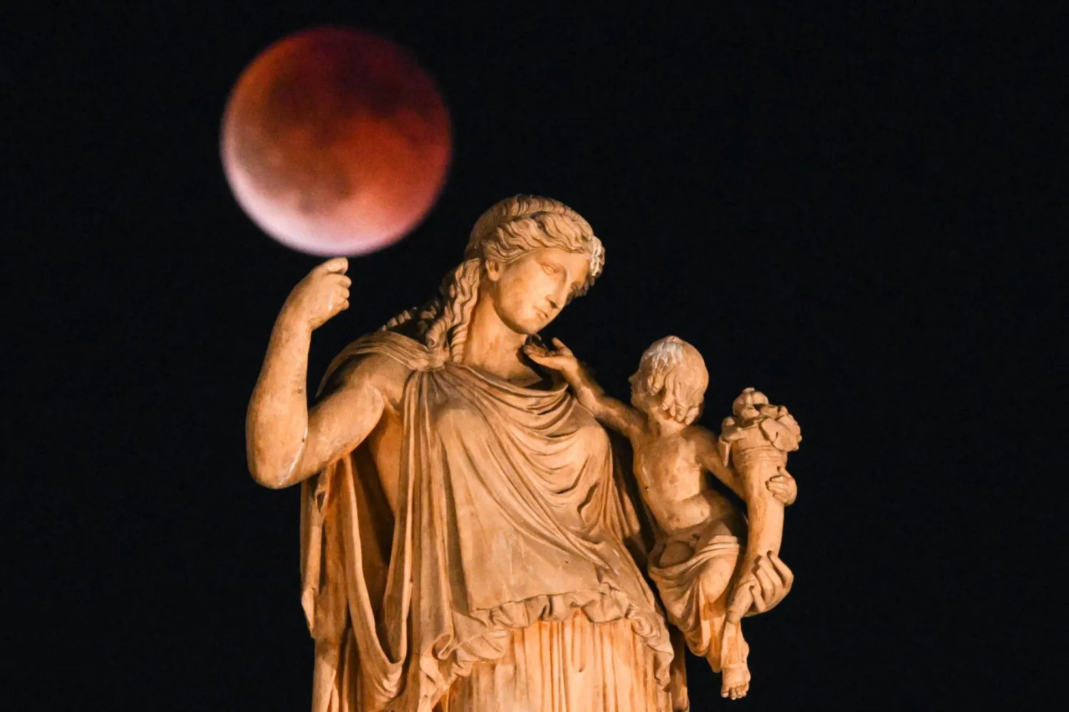 «القمر الدموي» أثناء خسوف القمر مع تمثال للإلهة اليونانية القديمة إيرين تحمل طفلاً في المقدمة (أ.ف.ب)