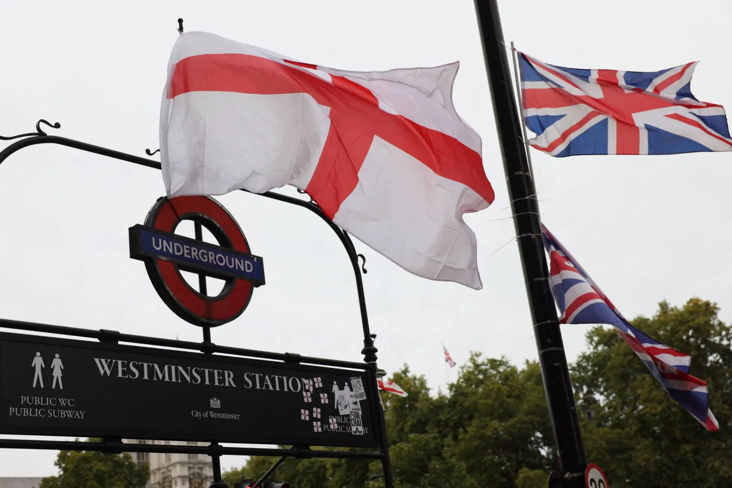 تتدلى أعلام العَلم البريطاني وصليب القديس جورج من لافتة فوق محطة مترو وستمنستر في اليوم التالي لمظاهرة مناهضة للهجرة نظمها الناشط البريطاني المناهض للهجرة ستيفن ياكسلي لينون المعروف أيضاً باسم تومي روبنسون بلندن 14 سبتمبر 2025 (رويترز)