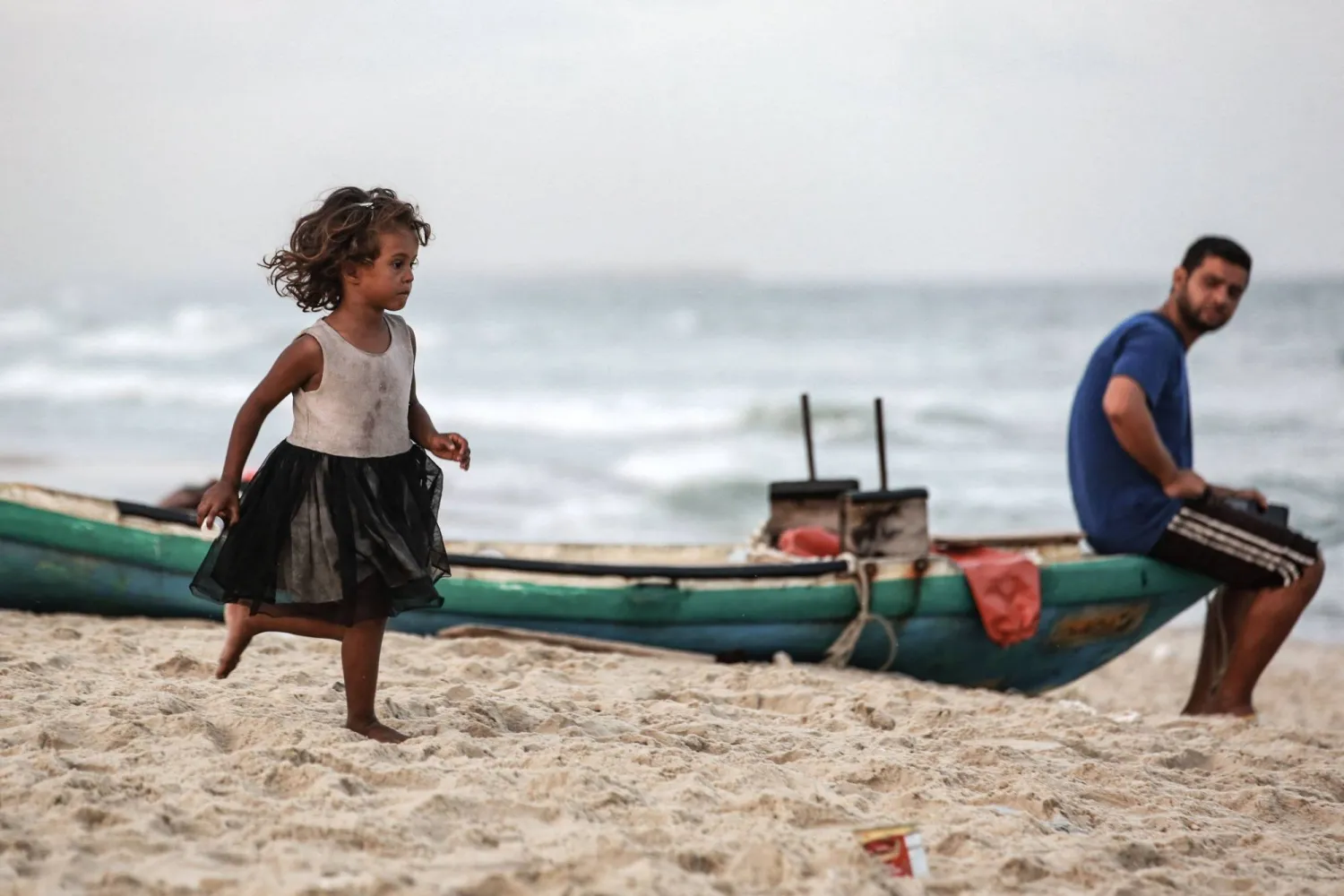 فتاة فلسطينية نازحة تركض على طول الشاطئ بينما يجلس رجل على متن قارب صيد في دير البلح وسط غزة يوم الأحد (أ.ف.ب)