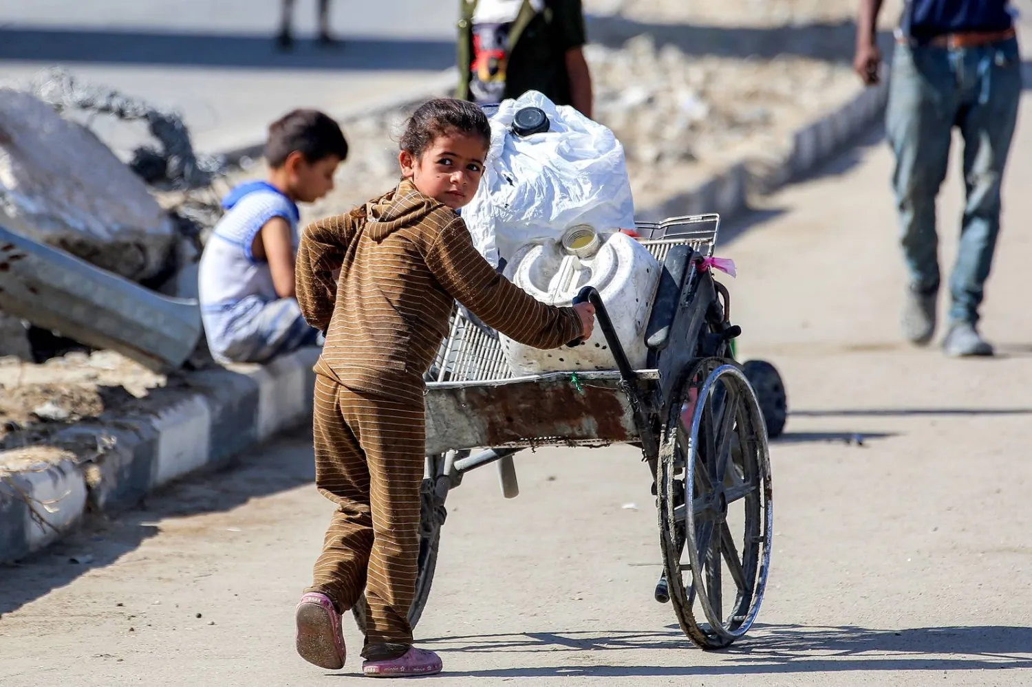 فتاة تدفع كرسياً متحركاً مكسوراً محملاً بصفائح المياه على طريق في مخيم للنازحين جراء الحرب شمال النصيرات (أ.ف.ب)