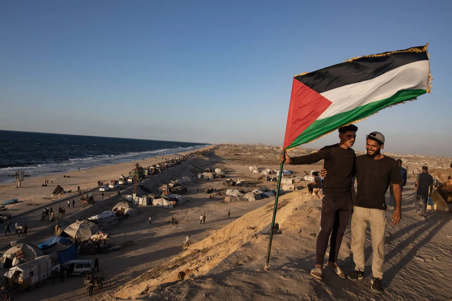 رجلان يحملان علم فلسطين بينما يسير مواطنون نازحون حاملين أمتعتهم على طول طريق الرشيد الساحلي بالقرب من وادي غزة في وسط قطاع غزة (إ.ب.أ)