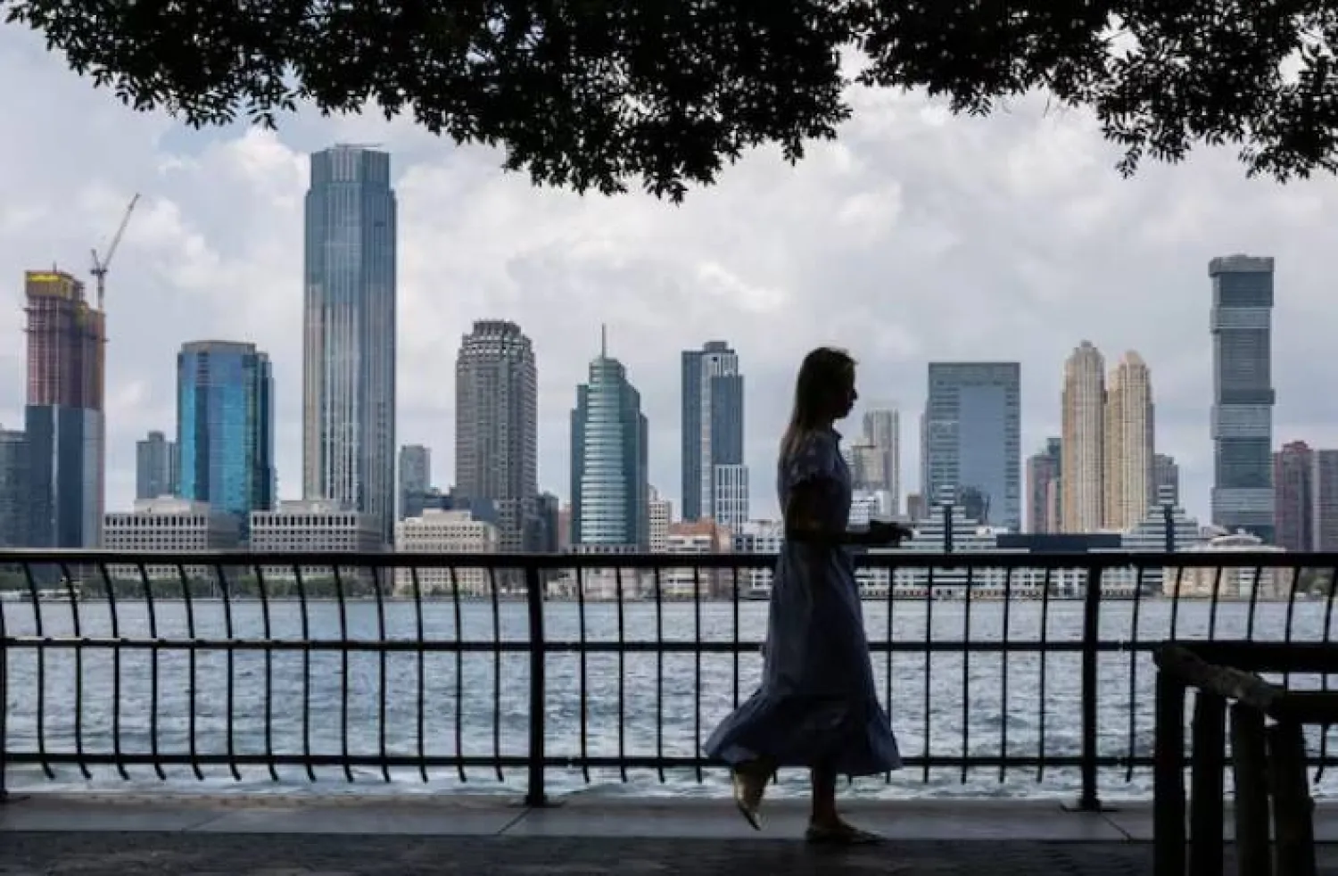 امرأة تمشي على طول نهر هدسون مع أفق جيرسي سيتي في الخلفية بمدينة نيويورك (رويترز)