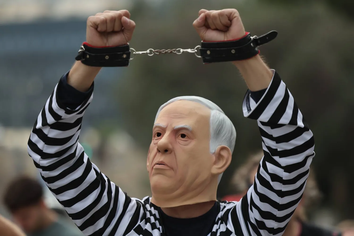 معارض لنتنياهو يضع قناعاً يمثله ويلبس زي سجين ويداه مكبلتان خارج مقر المحكمة بتل أبيب في ديسمبر 2024 (إ.ب.أ)