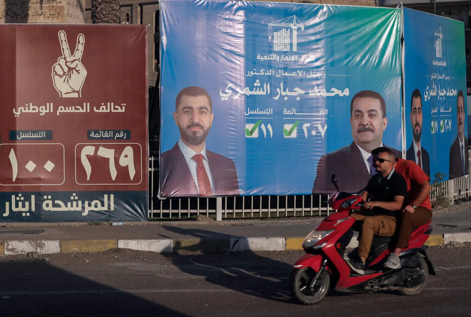 شخصان يقودان دراجتهما البخارية أمام لوحات إعلانية انتخابية في أحد شوارع بغداد (أ.ف.ب)